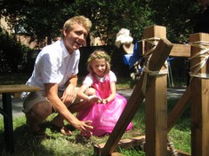 Ein Mann kniet mit seiner als Prinzessin verkleideten Tochter im Gras und spielt mit einem kleinen Katapult.