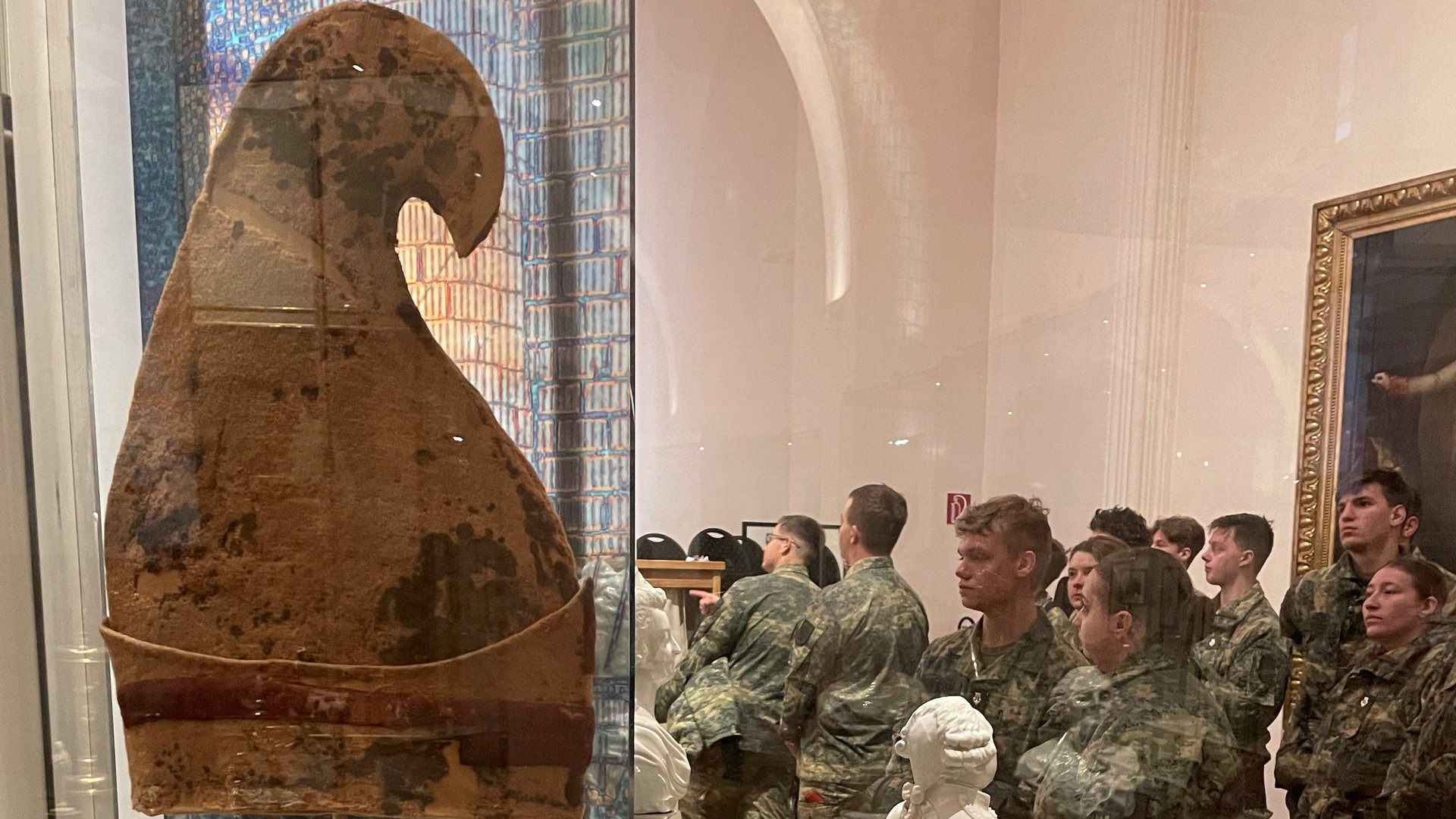 Das Bild zeigt eine Jakobinermütze, die in einer Vitrine ausgestellt ist. Die Jakobinermütze war ein Symbol der Bewegung der Französischen Revolution. Hinter der Vitrine sind gerahmte Lithographien an der Wand zu sehen, die möglicherweise historische Szenen der Revolution darstellen, sowie kleinere Statuetten.