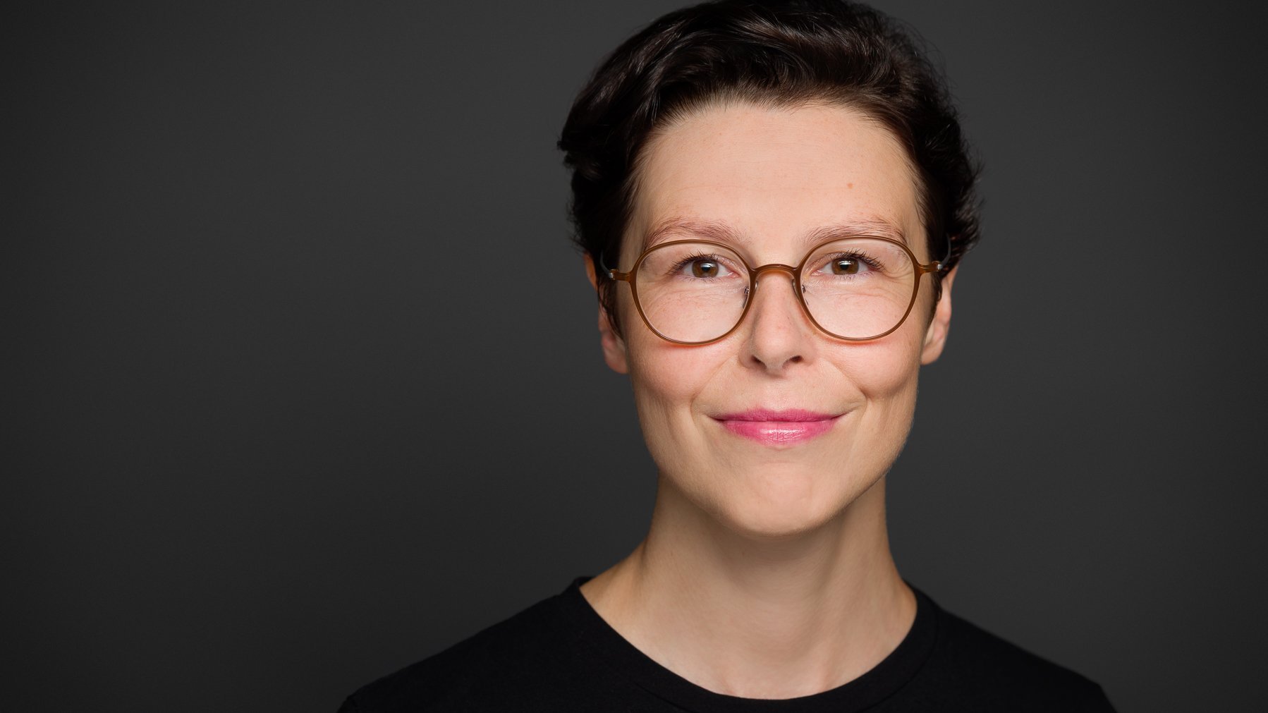 © © Fotografie Carolin Bohn 2023 Auf dem Foto ist die Militär- und Medizinhistorikerin Sabine Jesner zu sehen. Sie hat kurzes, dunkles Haar und trägt eine runde Brille mit einem hellen Rahmen. Sie trägt ein schwarzes Oberteil und lächelt leicht in die Kamera. Der Hintergrund ist einfarbig dunkelgrau, wodurch ihr Gesicht deutlich hervorsticht. Das Porträt ist professionell aufgenommen und zeigt sie in einer ruhigen und selbstbewussten Pose.