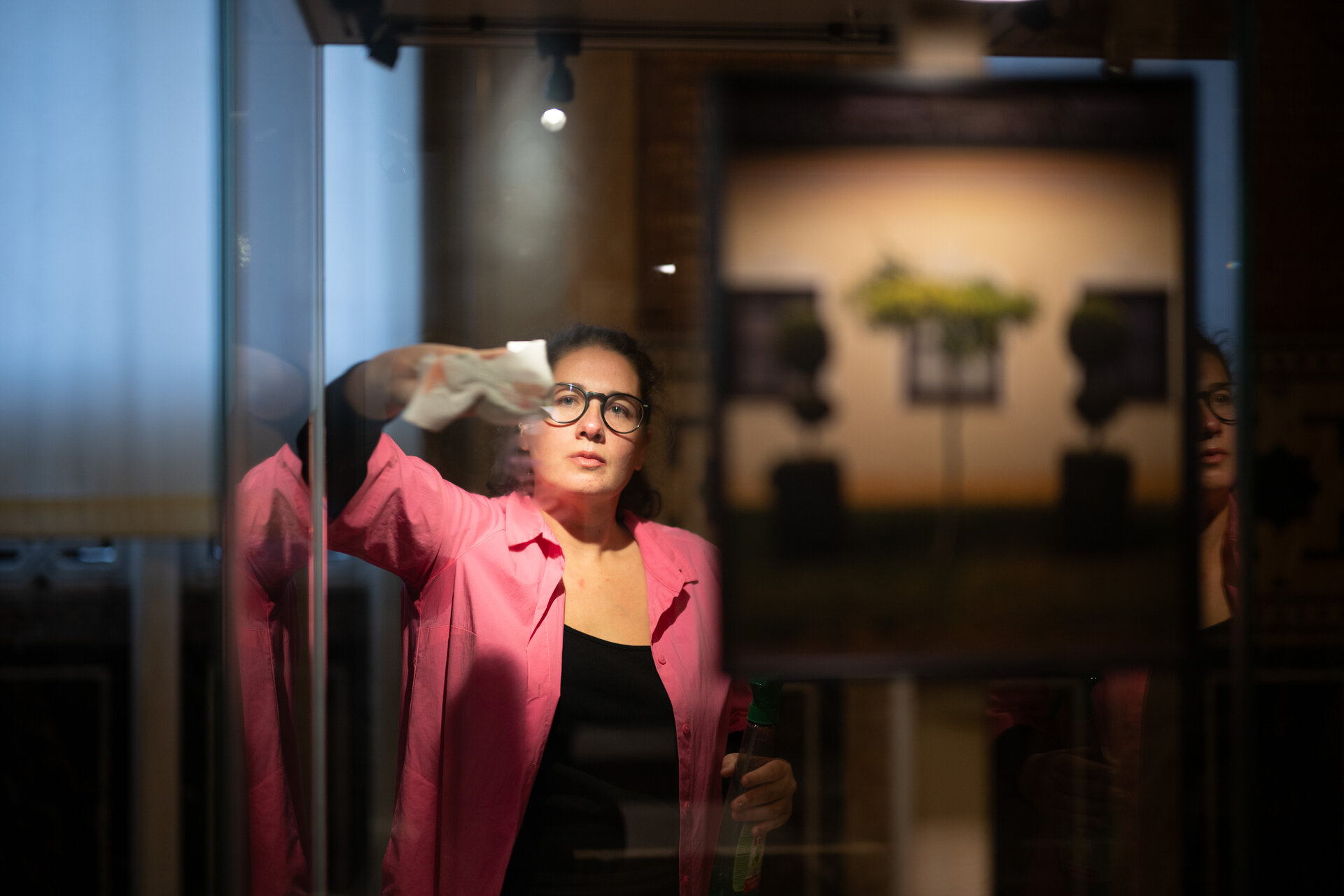 Im Bild sehen sie die Künstlerin Raffaela Bielesch wie sie eine Glasscheibe mit einem Tuch putzt. Der Hintergrund ist dunkel gehalten. Der Vordergrund ist mit hellem Licht beleuchtet. 