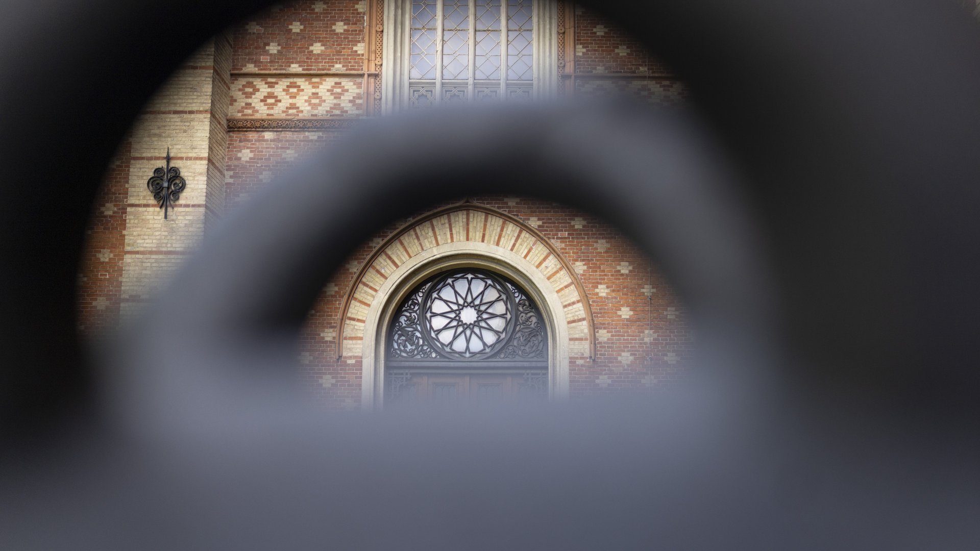 © BMLV/Daniel TRIPPOLT Auf diesem Bild der Außenfassade des HGMs ist eine kunstvoll gestaltete Tür zu sehen. Das Foto scheint durch ein Visier oder ein Rohr einer Kanone aufgenommen worden zu sein. Über der Tür ist ein halbrundes Fenster mit filigranen Gittermuster zu sehen. Das Fenster ist von einem steinernen Rahmen umgeben. Er ist mit roten und cremefarbenen Backsteinen verziert, es bildet ein Bogenmuster. Oberhalb des Fenster ist ein rechteckiges Fenster, das in mehrere Abschnitte unterteilt ist, mit einer aufwendigen Verglasung zu erkennen. Die umgebende Backsteinfassade ist in einem warmen Rotton gehalten und weist ein Muster aus helleren und dunkleren Steinen auf. Links neben der Tür ist ein dekoratives schmiedeeisernes Element an der Wand angebracht, das wie ein stilisierter Pfeil aussieht.