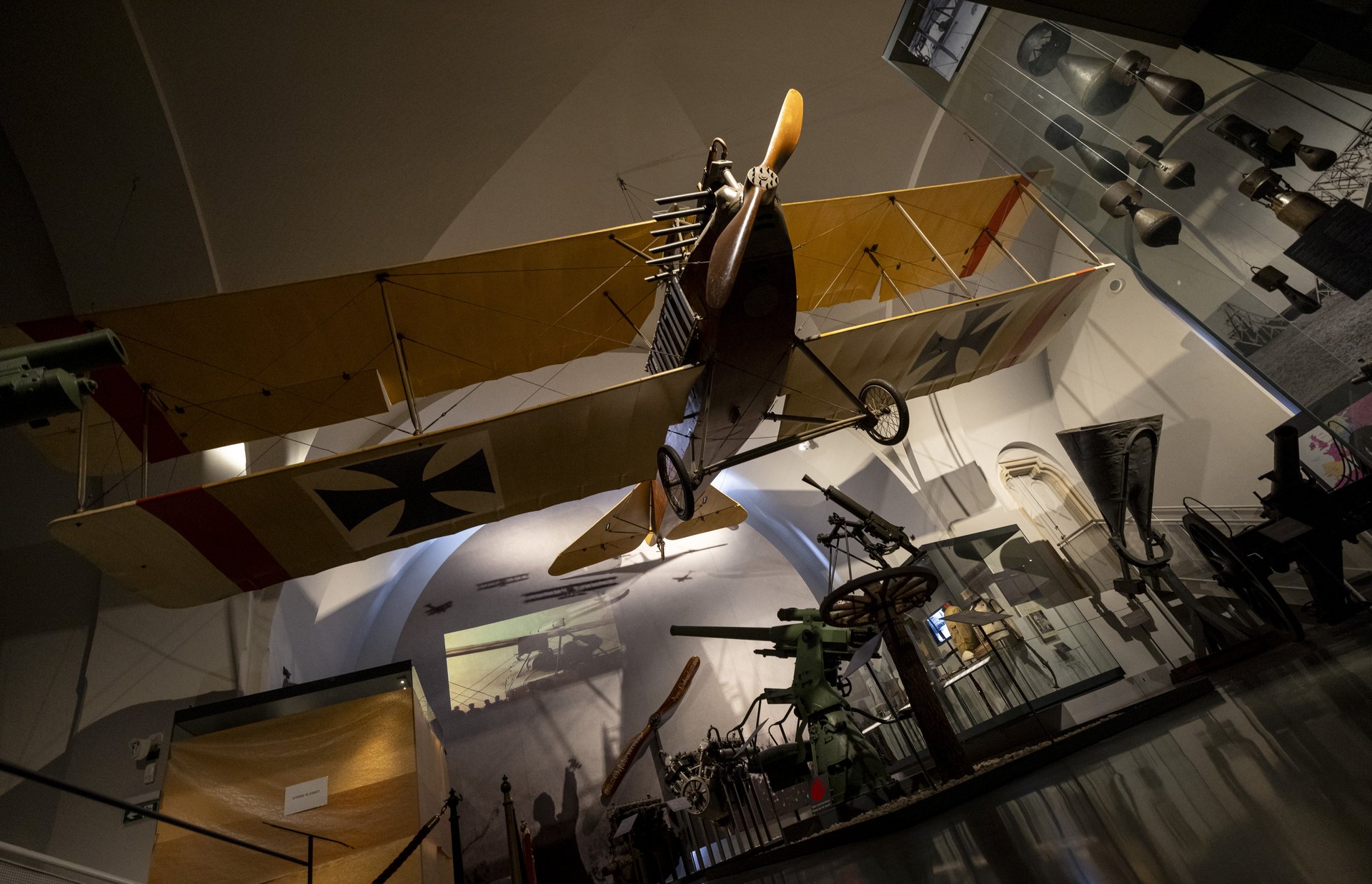 Dieses Bild zeigt ein frühes Militärflugzeug von der Decke hängend, es stammt aus der Zeit des ersten Weltkriegs, erkennbar an dem Eisernen Kreuz auf den Flügeln. Die umgebende Ausstellung umfasst verschiedene Artefakte und Ausrüstungsgegenstände wie Artilleriestücke, einen Motor und anderes militärisches Zubehör. Die Beleuchtung hebt das Flugzeug hervor und wirft Schatten an die Wände, wodurch eine eindrucksvolle Atmosphäre für die Besucher entsteht.