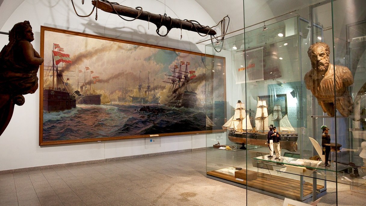 Das Bild präsentiert eine eindrucksvolle Ansammlung von Galionsfiguren in Vitrinen. In der Mitte des Raumes dominiert ein großes Gemälde, das Kriegsschiffe in Aktion zeigt und die maritime Geschichte lebendig werden lässt. Neben dem Gemälde steht ein detailgetreues Segelschiffmodell in einer weiteren Vitrine, welches die handwerkliche Meisterschaft und die Vielfalt der Schifffahrtstraditionen illustriert.
