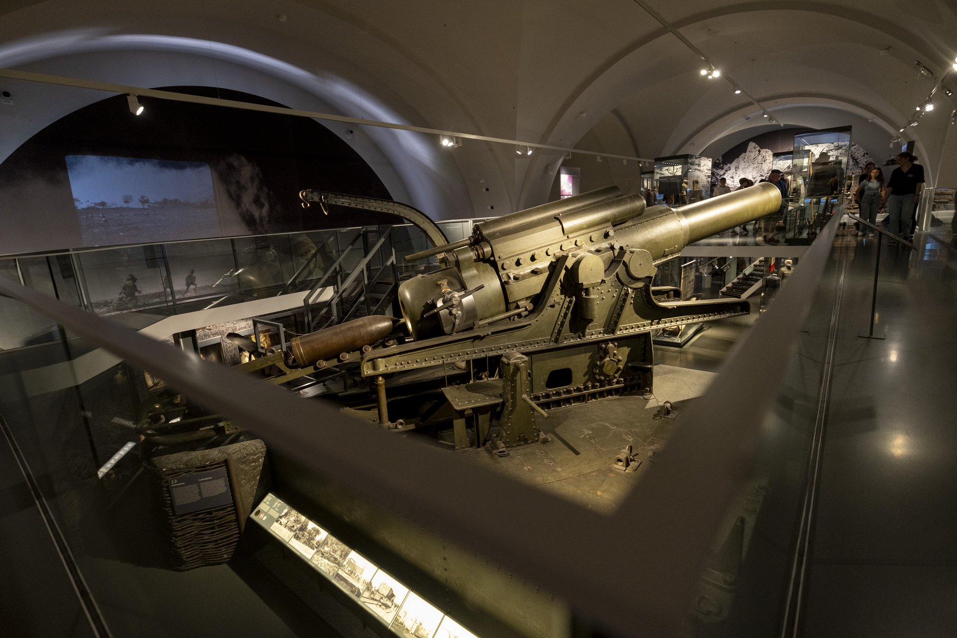  Im Zentrum der Aufnahme steht ein großes Geschütz, vermutlich ein schwerer Artilleriewaffen wie eine Haubitze oder Kanone, die während des Krieges eingesetzt wurde. Die Waffe ist detailliert dargestellt und in einem Zustand, der ihre Nutzung und Mechanik deutlich macht.  Im Hintergrund ist eine große Fotografie oder ein Filmprojektion zu sehen, die Soldaten im Einsatz zeigt, was die historische Bedeutung und den Einsatz der gezeigten Waffe unterstreicht. Besucher der Ausstellung sind im Hintergrund erkennbar, die sich die verschiedenen Exponate ansehen. Die Ausstellungshalle ist groß und gut beleuchtet, was eine eindrucksvolle und immersive Präsentation der historischen Artefakte ermöglicht. Informationstafeln und weitere Ausstellungsstücke sind ebenfalls sichtbar und bieten zusätzliche Kontexte und Details zur gezeigten Artillerie und ihrer Rolle im Ersten Weltkrieg.