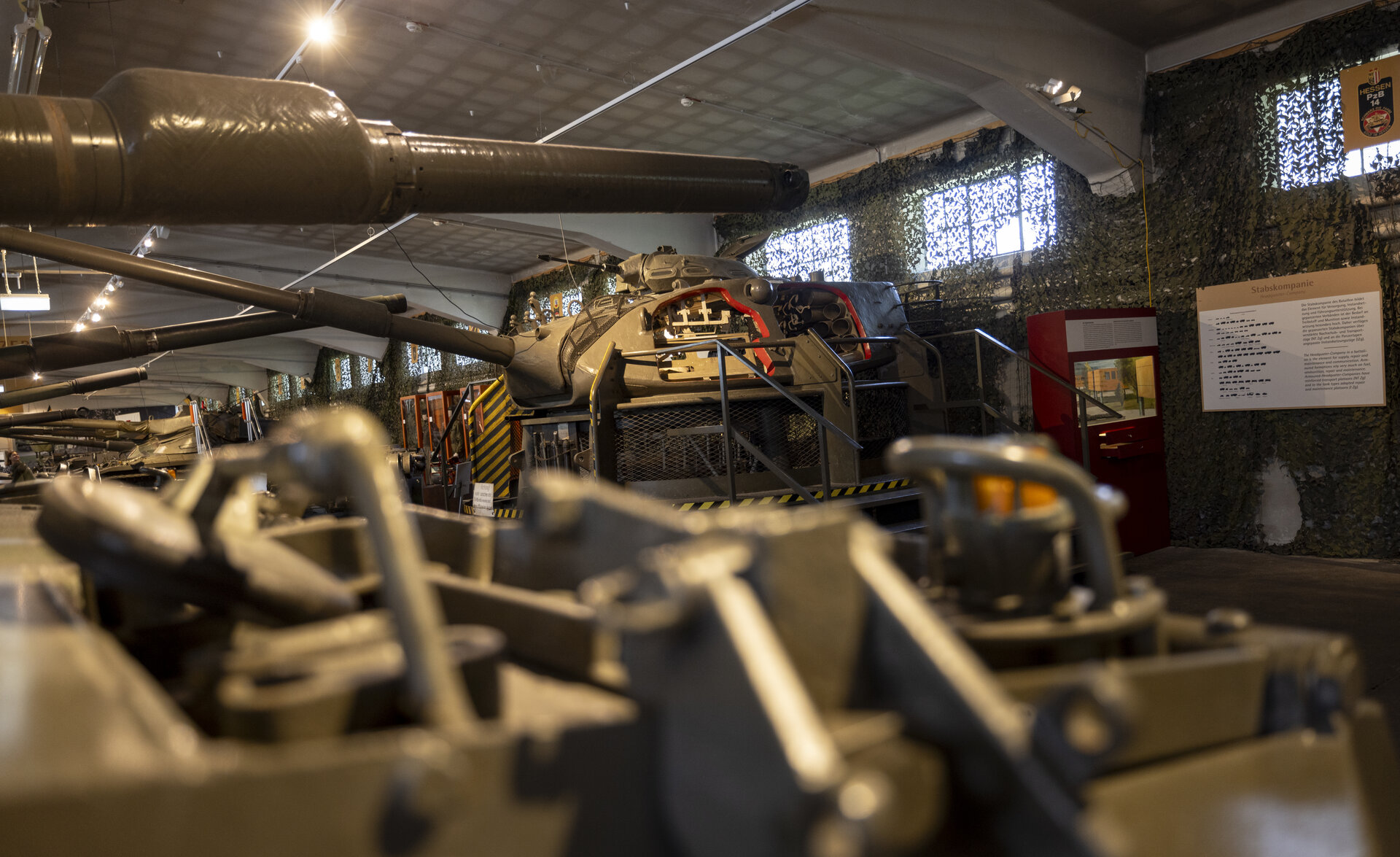 Ein Bild der Panzerhalle im Heeresgeschichtlichen Museum. Im Vordergrund sind die Kanonenrohre mehrerer Panzer zu sehen, die diagonal ins Bild ragen. In der Mitte des Bildes steht ein großer, gut beleuchteter Panzer, teilweise von Sicherheitsgeländern umgeben. Die Halle ist mit Tarnnetzen an den Wänden dekoriert, durch die Licht von kleinen Fenstern hereinfällt. Auf der rechten Seite befindet sich eine Informationstafel mit historischen Details. Die Szenerie ist von einem warmen Licht durchflutet, das die militärischen Ausstellungsstücke hervorhebt.