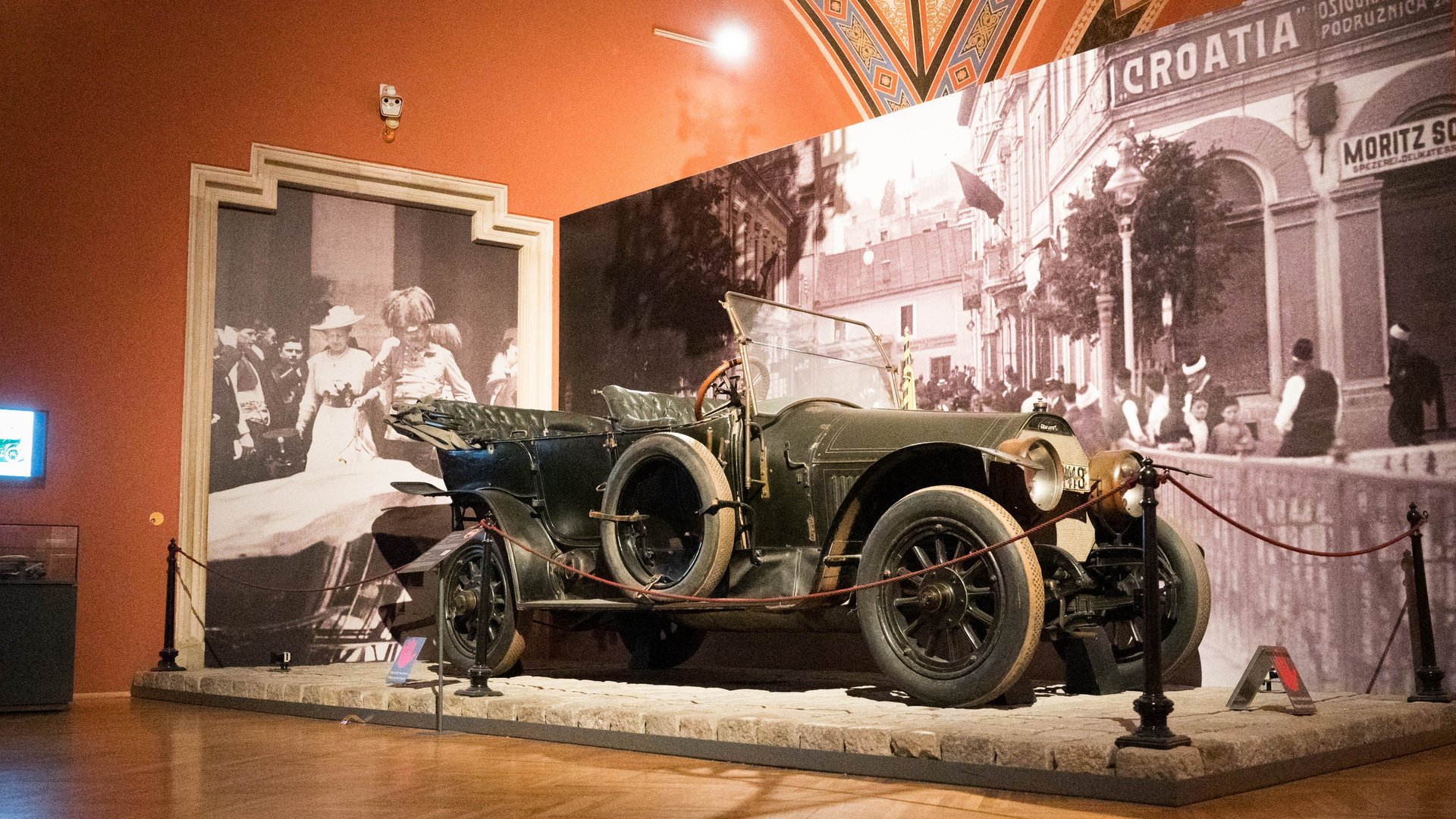 © RONJA_KLIMA/HBF Ein historisches Automobil, ein Gräf & Stift, ausgestellt im Saal "Das Attentat von Sarajevo" des Heeresgeschichtlichen Museums. Das Fahrzeug ist mit einer Absperrung umgeben und auf einem Podest platziert. Im Hintergrund sind großformatige Schwarz-Weiß-Fotografien zu sehen, die Szenen aus der Zeit des Attentats in Sarajevo zeigen. Eine Fotografie zeigt Menschen in historischen Kleidern, eine andere eine Straßenszene mit Gebäuden und dem Schriftzug "CROATIA". Der Ausstellungsraum hat eine warme Beleuchtung und eine verzierte Decke.