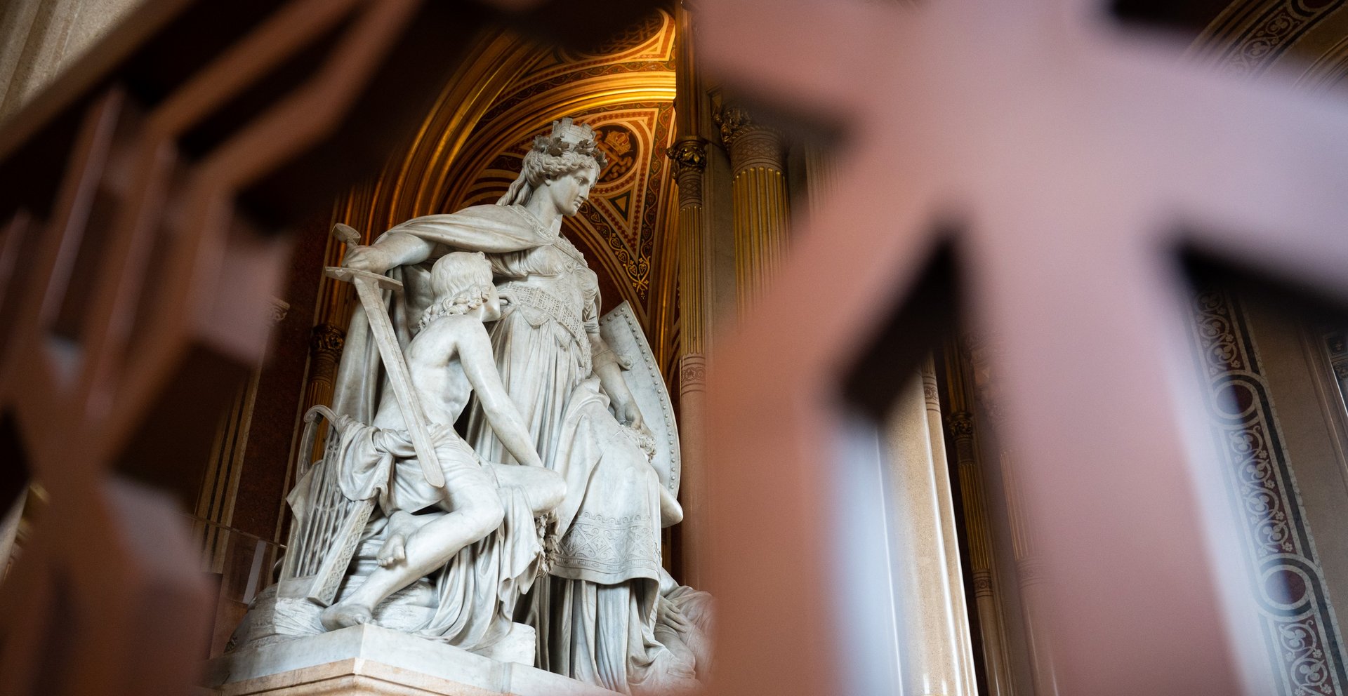 Das Bild zeigt einen Teil der Feldherrenhalle im Heeresgeschichtlichen Museum, die den Aufgang zur Ruhmeshalle darstellt. Durch ein dekoratives Gitterwerk im Vordergrund blickt man auf eine beeindruckende Marmorskulpturengruppe. Die Skulptur zeigt eine majestätische Figur einer Frau in antiker Rüstung, die eine schützende Pose einnimmt und einen Schild hält. Neben ihr ist eine weitere Figur eines Kriegers, der zu ihr aufblickt. Die kunstvolle Gestaltung der Figuren und die detaillierten Verzierungen der Kleidung und Ausrüstung sind deutlich erkennbar.