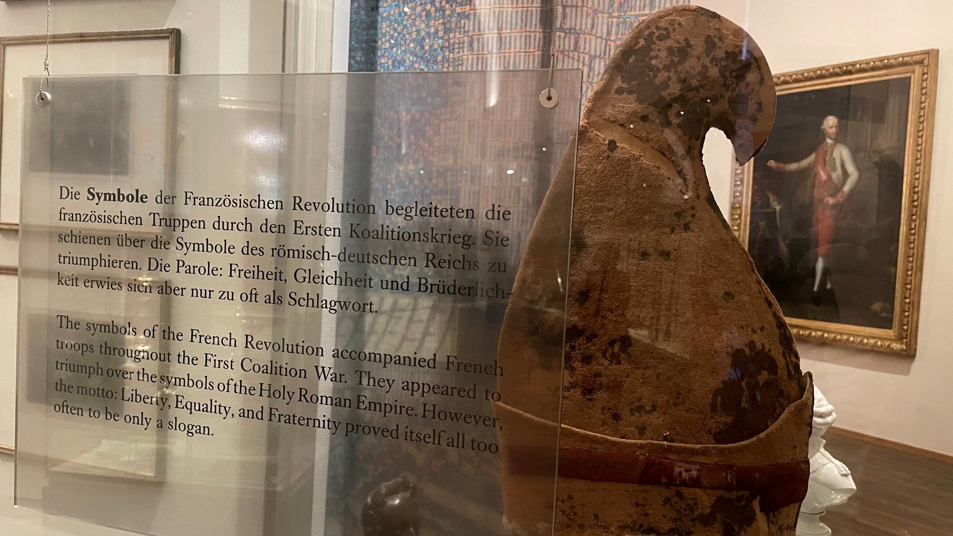 Das Bild zeigt eine Jakobinermütze, die in einer Vitrine ausgestellt ist. Die Jakobinermütze war ein Symbol der Bewegung der Französischen Revolution. Hinter der Vitrine sind gerahmte Lithographien an der Wand zu sehen, die möglicherweise historische Szenen der Revolution darstellen, sowie kleinere Statuetten.