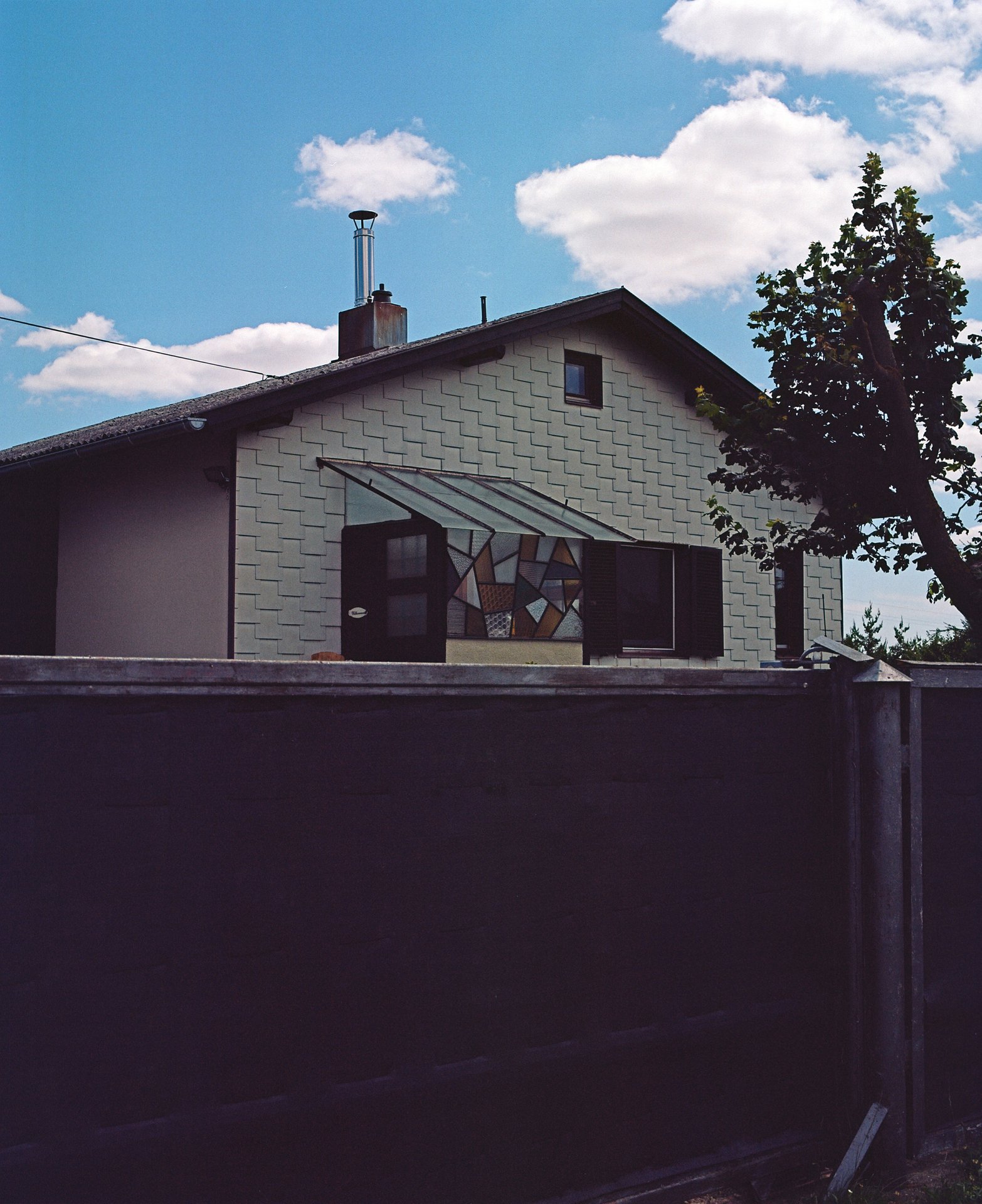 Auf diesem Bild ist ein Haus mit weißen Fassadenplatten verziert zu sehen, davor steht ein dunkelgehaltener Zaun. Der Himmel im Hintergrund ist leicht bewölkt und hellblau. Auf dem Dach des Hauses ragt ein Rauchfang mit einem Rohr empor. 