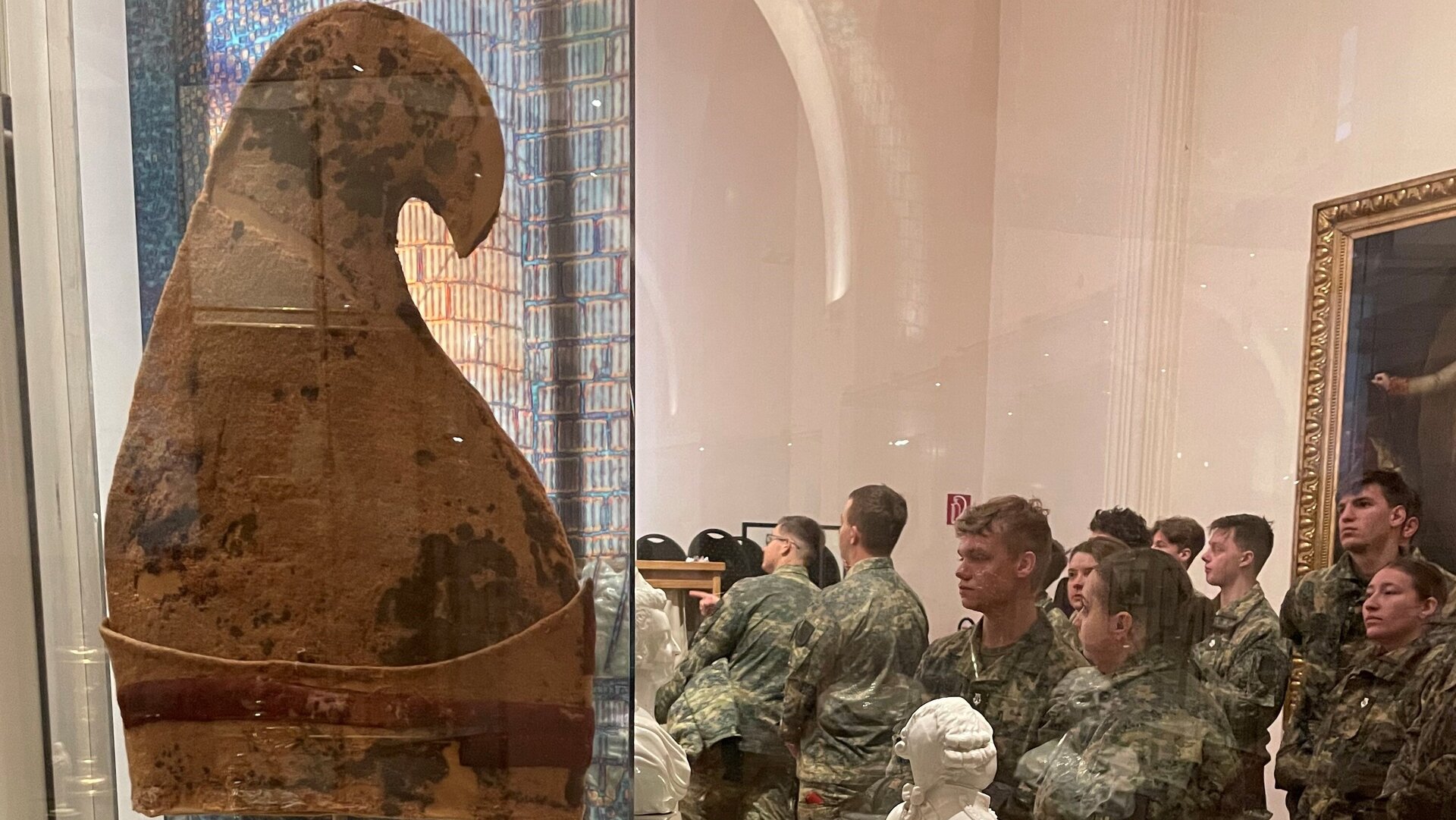 Das Bild zeigt eine Jakobinermütze, die in einer Vitrine ausgestellt ist. Die Jakobinermütze war ein Symbol der Bewegung der Französischen Revolution. Hinter der Vitrine sind gerahmte Lithographien an der Wand zu sehen, die möglicherweise historische Szenen der Revolution darstellen, sowie kleinere Statuetten. 