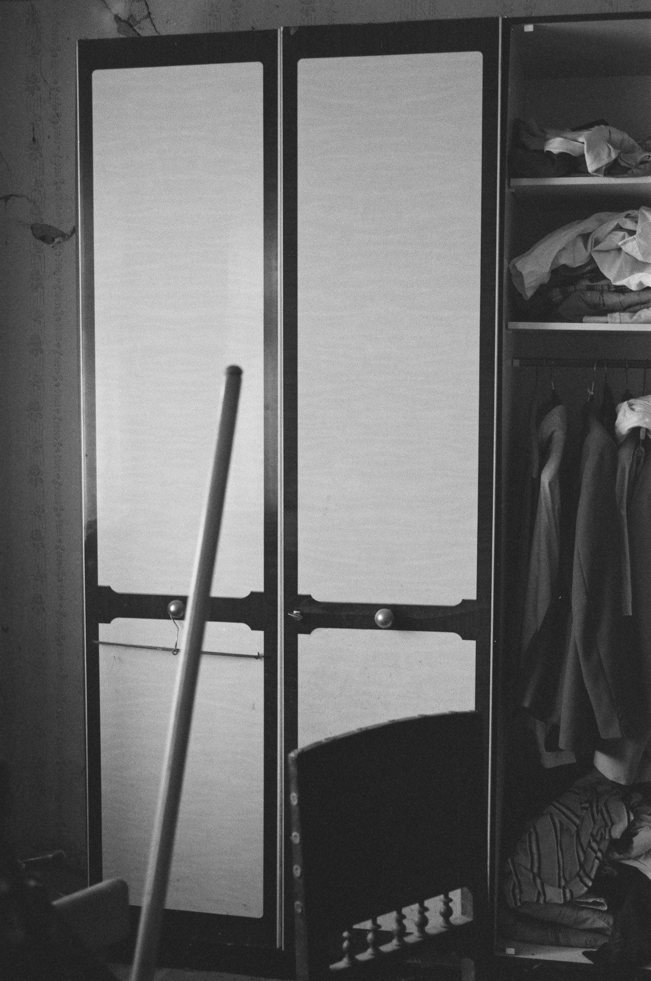 Auf diesem schwarz weiß gehaltenen Bild sieht man einen weißen Kleiderschrank, wessen Türen nur angelehnt zu sein scheint. Vor dem Kleiderschank sieht rechts den Kopfstück eines Bettes. Links auf dem Bild sieht man einen Besenstil. Rechts neben dem Kleiderschrank steht ein weiterer Kleiderschrank, in denen sich zusammengelegtes Gewand befindet. 