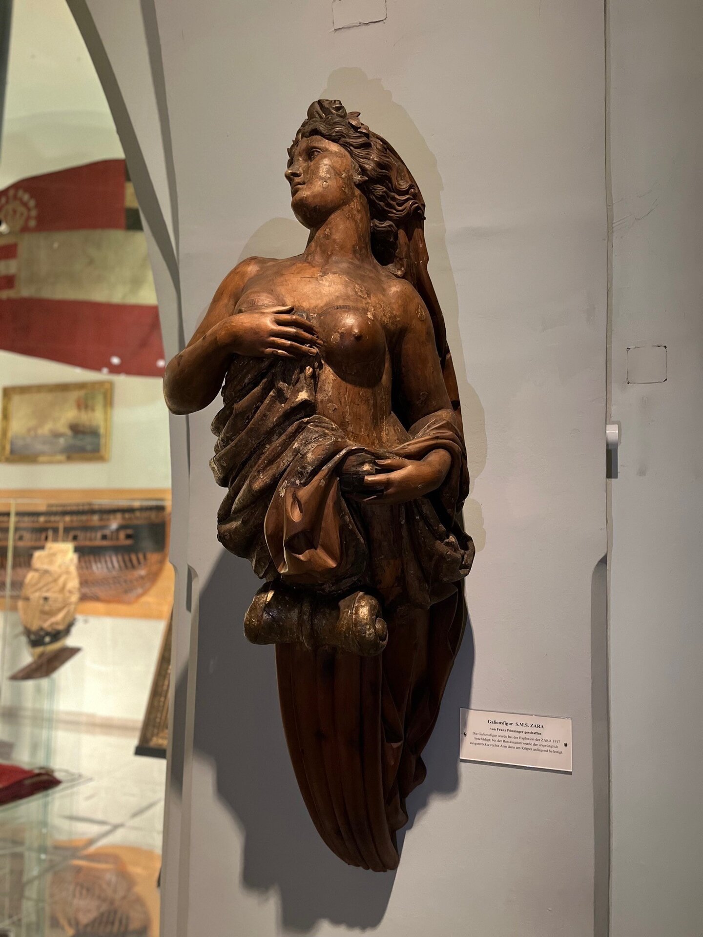 Zu sehen ist die hölzerne Galionsfigur der SMS Zara, die eine weibliche Gestalt mit freiem Oberkörper und seitlich abgewandtem Blick zeigt.