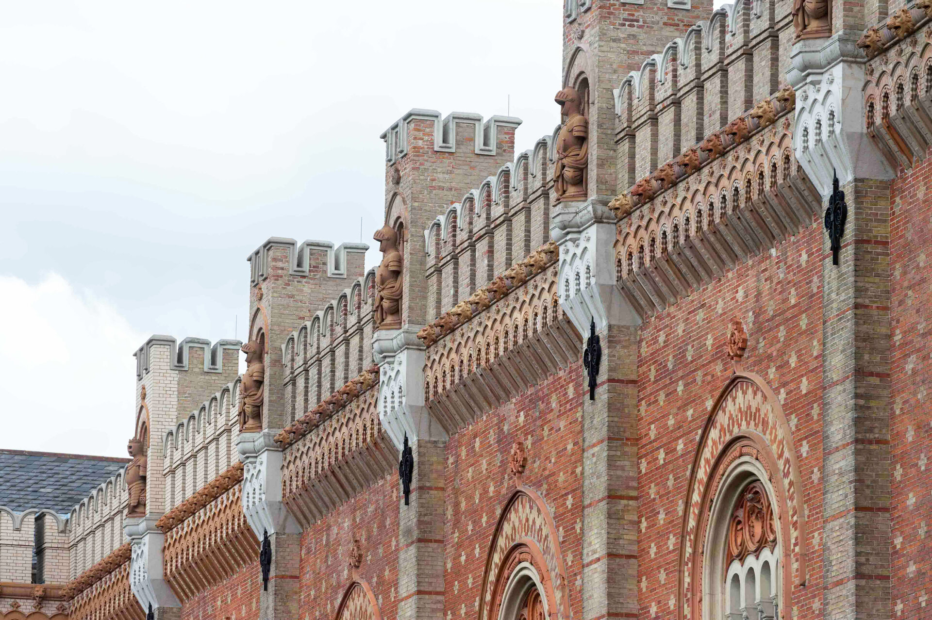 Das Bild zeigt die Außenfassade des Heeresgeschichtlichen Museums. Die Fassade ist aus roten und beigen Backsteinen gebaut und weist zahlreiche architektonische Details auf. Auffällig sind die Statuen von Rittern in Rüstungen, die in regelmäßigen Abständen entlang der Fassade in Nischen stehen. Über den Nischen und Fenstern sind Zinnen angebracht, die an mittelalterliche Burgen erinnern. Unter den Zinnen verlaufen dekorative Bögen mit detaillierten Mustern. 