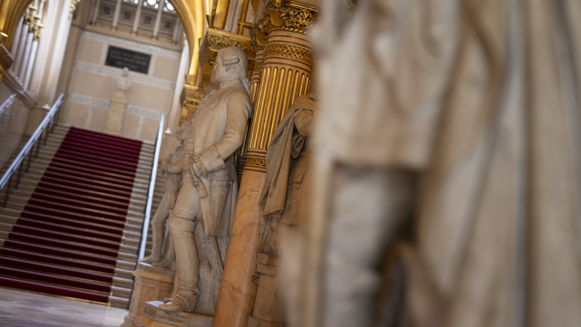 Auf dem Foto ist ein unschwarfes Detail einer Statue zu sehen, im Hintergrund sieht man jedoch die schärferen Details einer weiteren Statue sowie einen Treppenaufgang mit einem roten Teppich. 