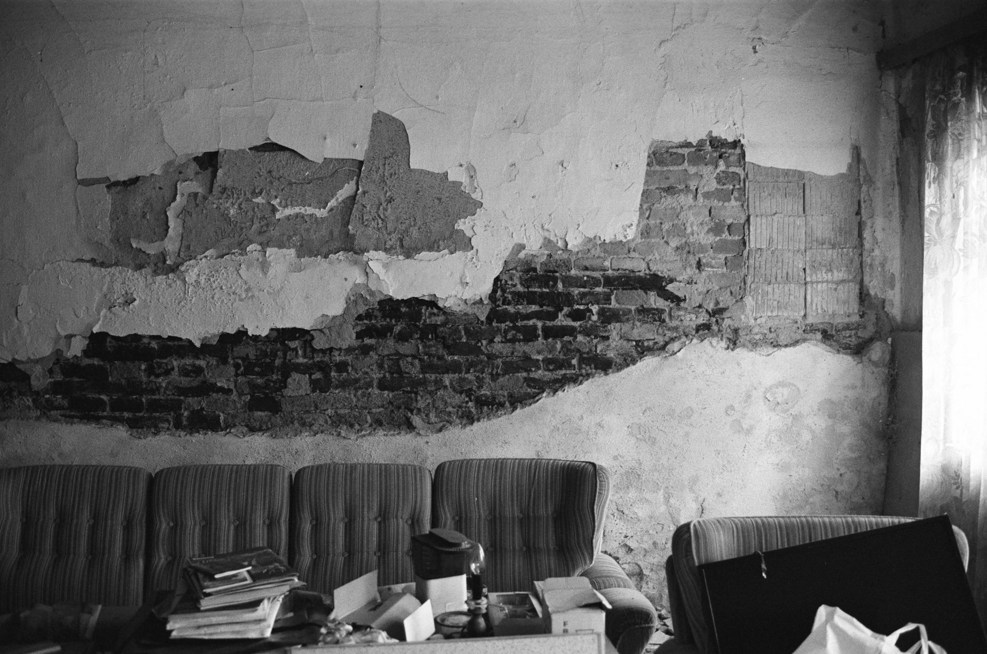 Auf diesem Bild sieht man eine aufgerissene Hausmauer, wessen Fassade runtergebröckelt ist. Hinter der Fassade sieht man das Ziegelmauerwerk. Vor der Mauer steht ein altes Sofa. Das Bild ist schwarz-weiß gehalten. 