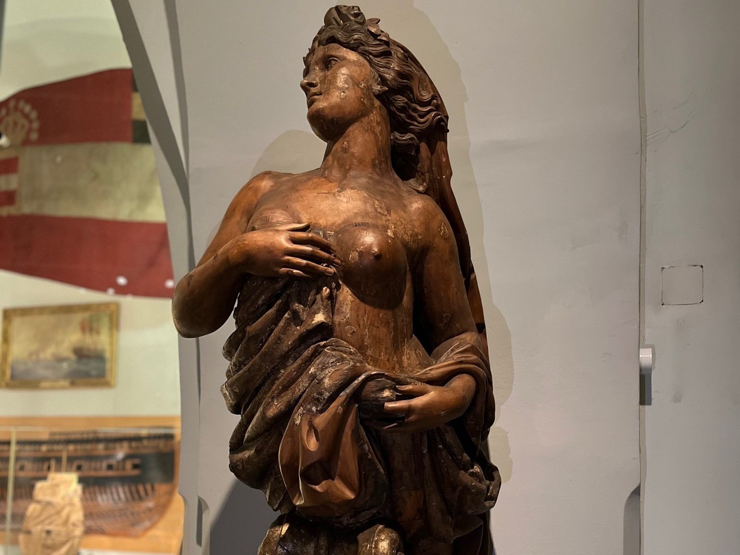 Zu sehen ist die hölzerne Galionsfigur der SMS Zara, die eine weibliche Gestalt mit freiem Oberkörper und seitlich abgewandtem Blick zeigt.
