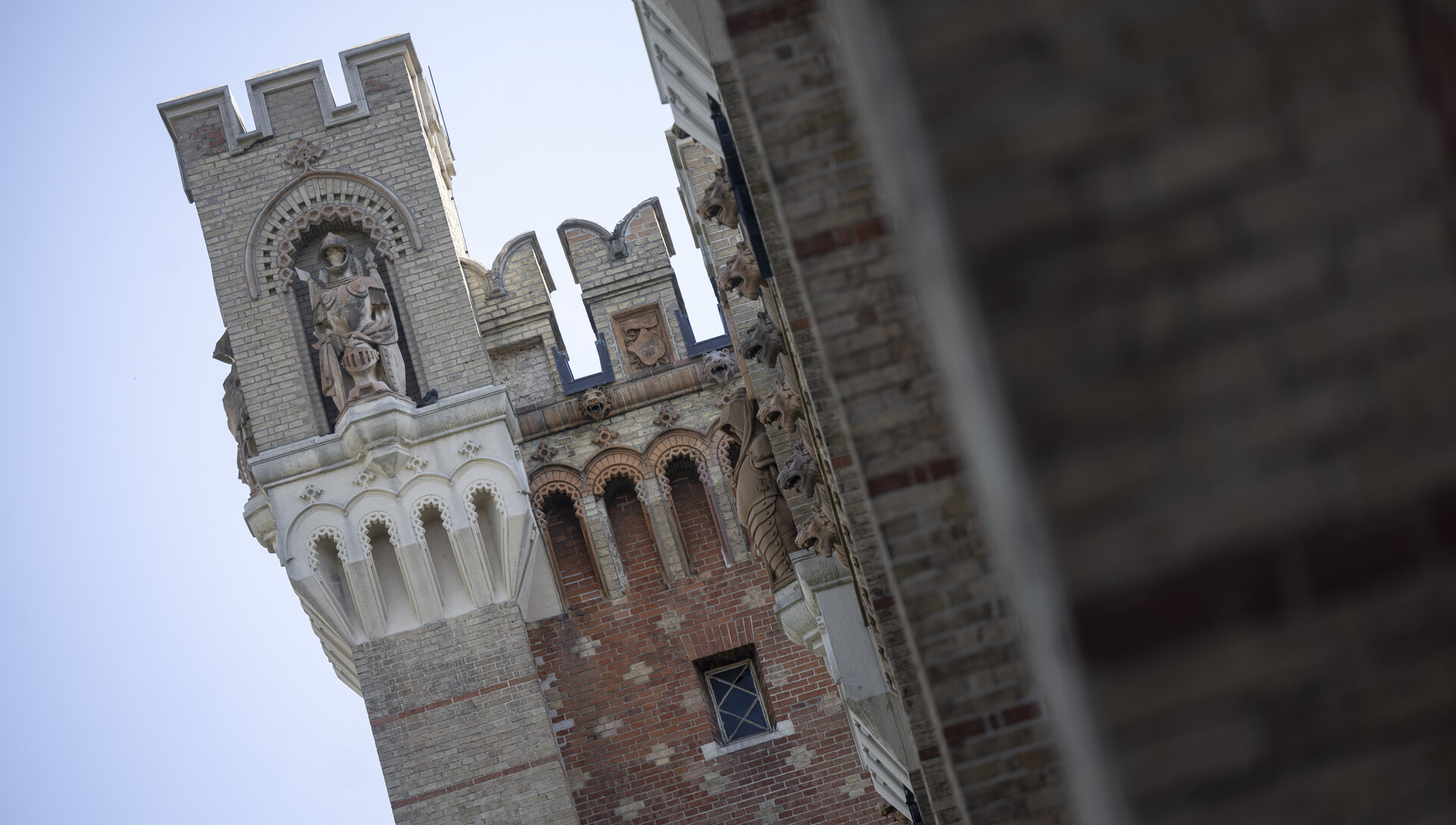 Dieses Bild zeigt ein Teil der Außenfassade des HGMs. Die Fassade zeigt eine Mischung aus Backstein- und Steinelementen. In der Mitte des Bildes befindet sich ein Turm mit einer dekorativen Aussparung, in der eine Ritterstatue steht. Der Ritter trägt ein Schild. Oberhalb der Statue ist ein Bogen mit Mustern zu erkennen. Der Turm hat eine gezackte Zierleiste, die dem mittelalterlichen Design nachgestellt ist. Neben dem Turm sind weitere Gebäudeteile zu sehen, die ebenfalls reich mit Verzierungen und Reliefs ausgestattet sind. Der Himmel ist klar und blau. 