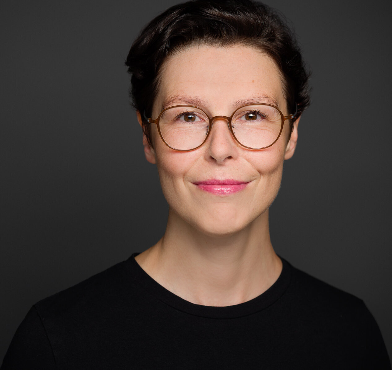 Auf dem Foto ist die Militär- und Medizinhistorikerin Sabine Jesner zu sehen. Sie hat kurzes, dunkles Haar und trägt eine runde Brille mit einem hellen Rahmen. Sie trägt ein schwarzes Oberteil und lächelt leicht in die Kamera. Der Hintergrund ist einfarbig dunkelgrau, wodurch ihr Gesicht deutlich hervorsticht. Das Porträt ist professionell aufgenommen und zeigt sie in einer ruhigen und selbstbewussten Pose.