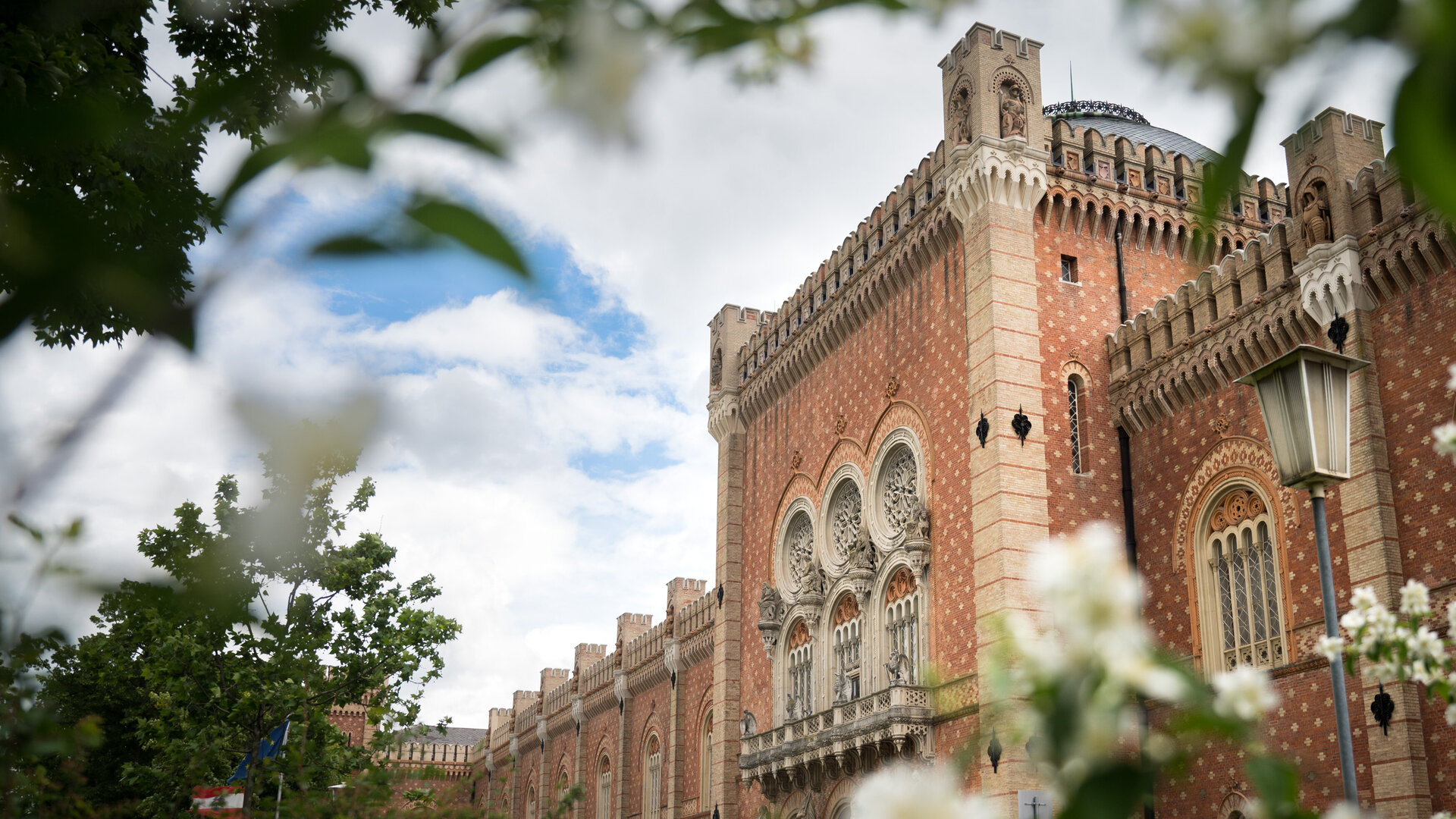  Durch Zweige voller blühender Blüten im Frühling hindurch blickt man auf die Seitenansicht des Heeresgeschichtlichen Museums. Deutlich sichtbar ist die ziegelrote Fassade mit den Fensterrosen, die mit unterschiedlichen Statuen geschmückt sind. Auch das Kuppeldach ist zu erkennen. Der Himmel ist blau und mit Wolken bedeckt.
