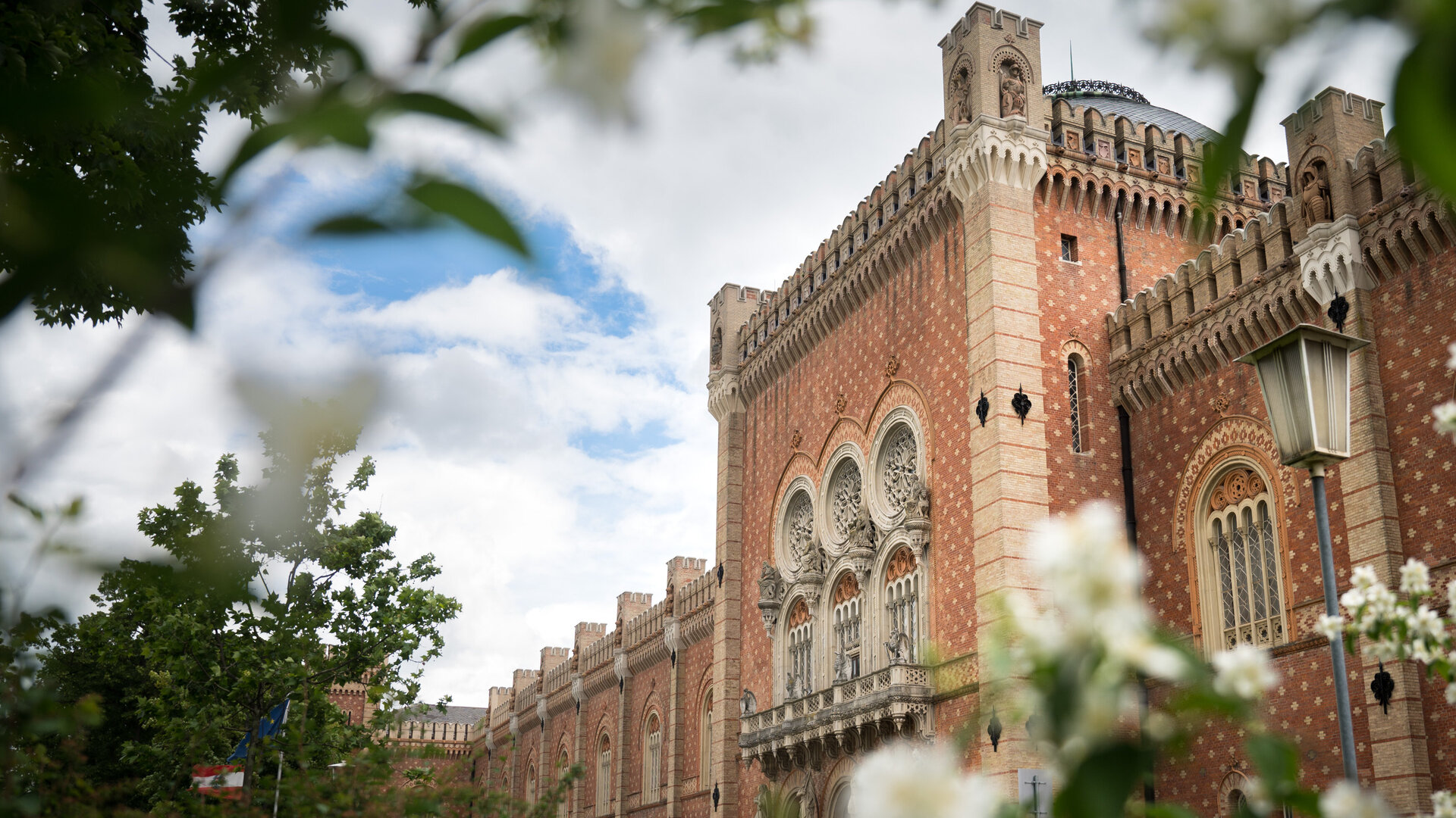  Durch Zweige voller blühender Blüten im Frühling hindurch blickt man auf die Seitenansicht des Heeresgeschichtlichen Museums. Deutlich sichtbar ist die ziegelrote Fassade mit den Fensterrosen, die mit unterschiedlichen Statuen geschmückt sind. Auch das Kuppeldach ist zu erkennen. Der Himmel ist blau und mit Wolken bedeckt.