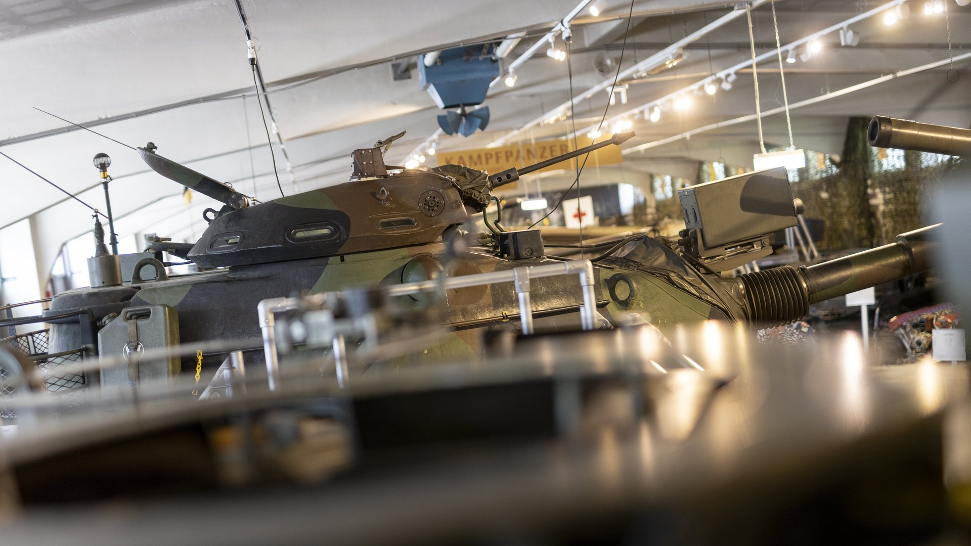 Ein Bild aus der Panzerhalle des Heeresgeschichtlichen Museums. Im Mittelpunkt des Bildes steht ein großer, olivgrüner Panzer, dessen Kanonenrohr nach rechts ragt. Das Fahrzeug ist mit zahlreichen Antennen und Ausrüstungsgegenständen versehen. Im Hintergrund sind weitere militärische Fahrzeuge und Ausrüstungen zu erkennen. Über dem Panzer hängt ein Schild mit der Aufschrift "KAMPFPANZER". 