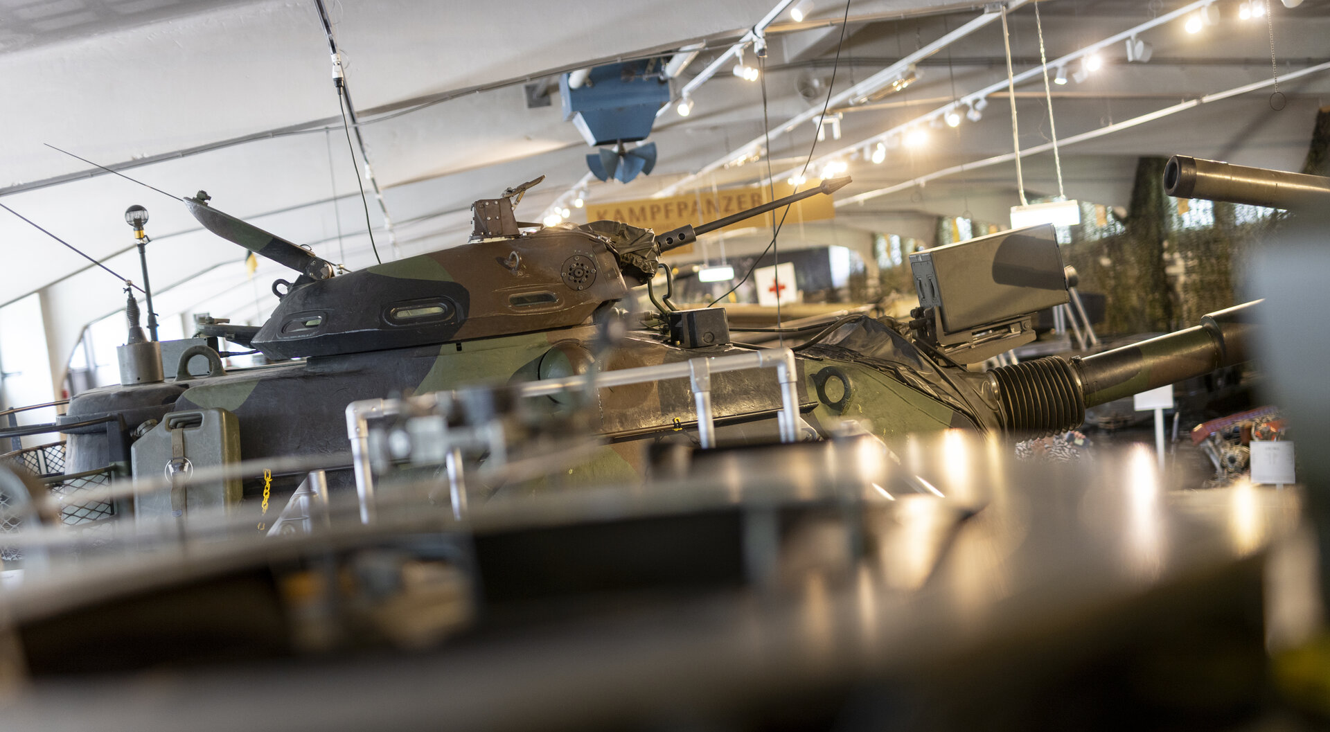 Ein Bild aus der Panzerhalle des Heeresgeschichtlichen Museums. Im Mittelpunkt des Bildes steht ein großer, olivgrüner Panzer, dessen Kanonenrohr nach rechts ragt. Das Fahrzeug ist mit zahlreichen Antennen und Ausrüstungsgegenständen versehen. Im Hintergrund sind weitere militärische Fahrzeuge und Ausrüstungen zu erkennen. Über dem Panzer hängt ein Schild mit der Aufschrift "KAMPFPANZER". 