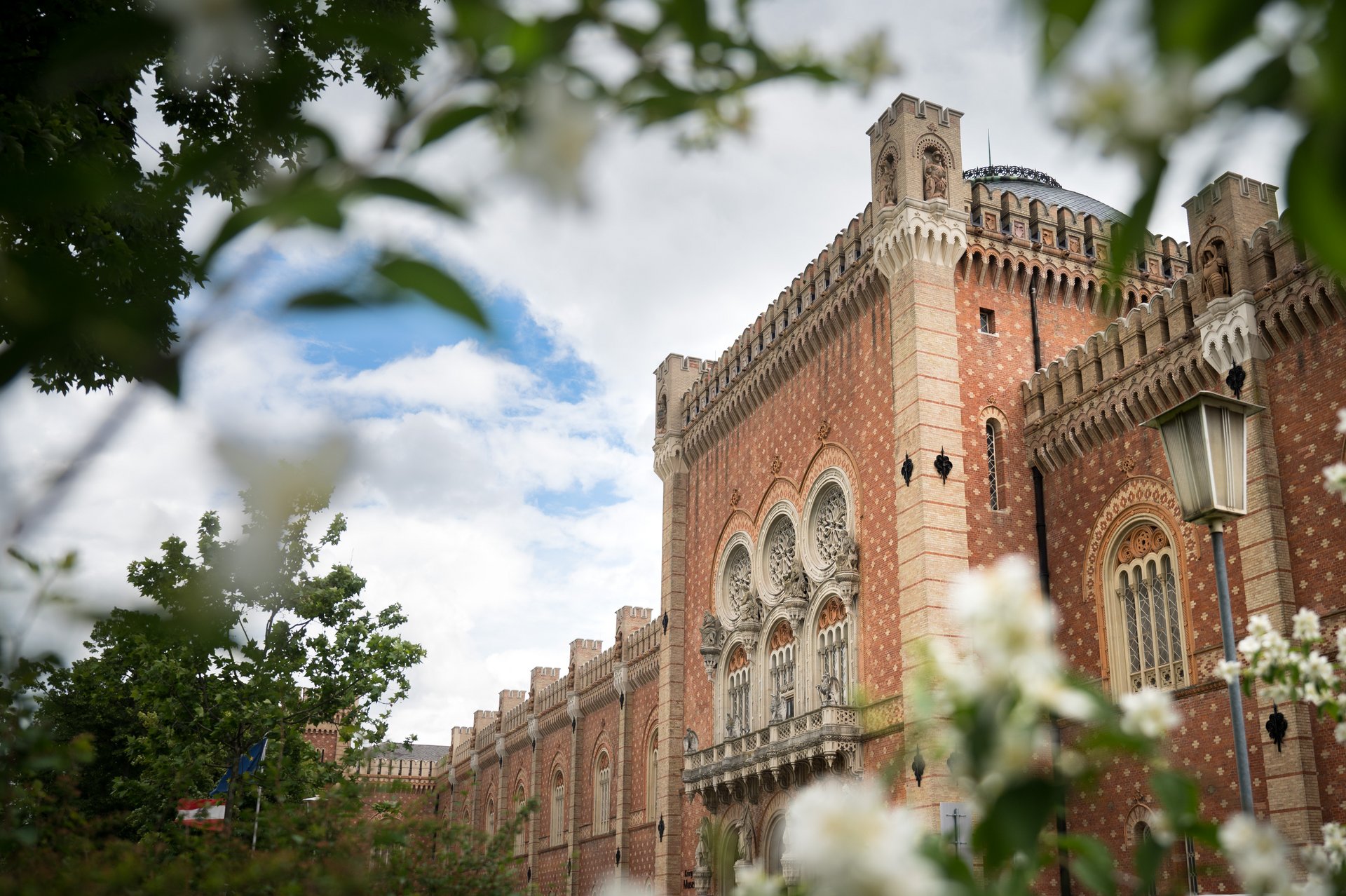  Durch Zweige voller blühender Blüten im Frühling hindurch blickt man auf die Seitenansicht des Heeresgeschichtlichen Museums. Deutlich sichtbar ist die ziegelrote Fassade mit den Fensterrosen, die mit unterschiedlichen Statuen geschmückt sind. Auch das Kuppeldach ist zu erkennen. Der Himmel ist blau und mit Wolken bedeckt.