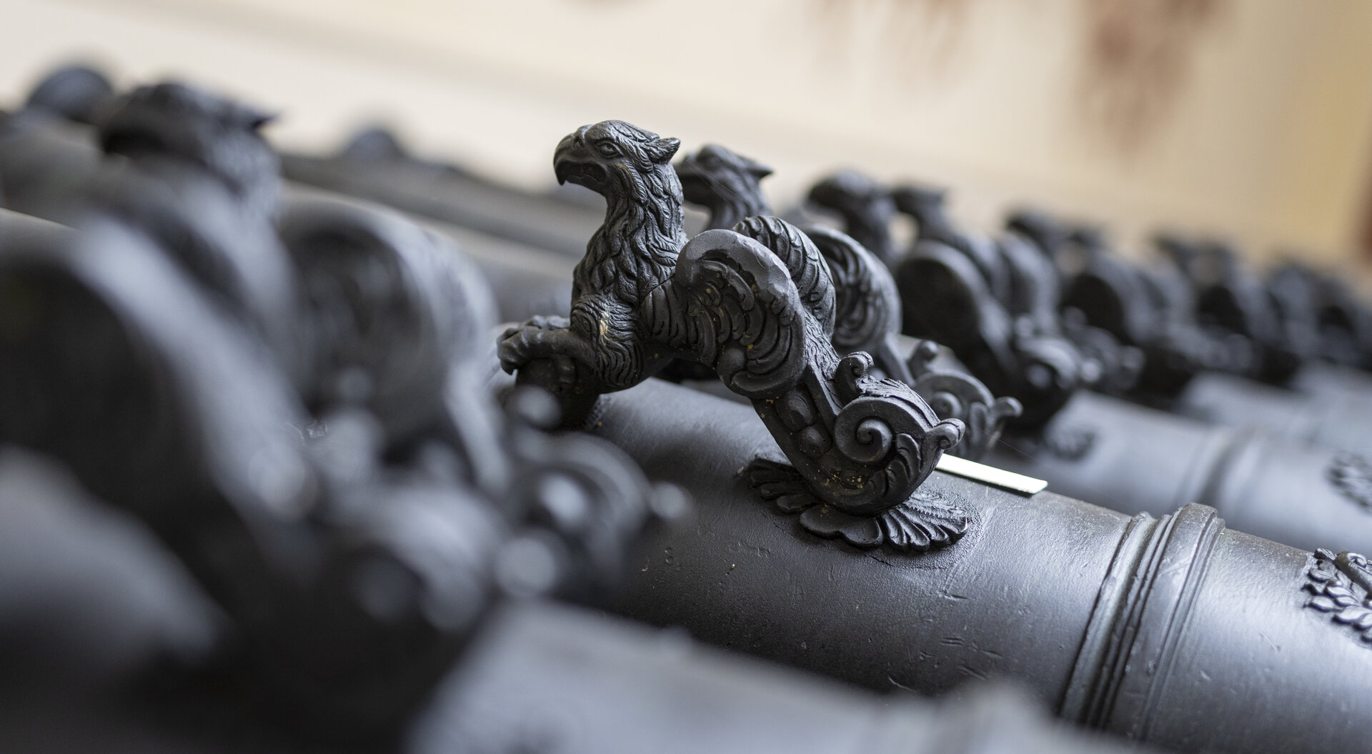 Auf dem Bild sind mehrere Geschützrohre in Rang und Glied aufgestellt, mit Blick fokussiert auf das kunstvoll dekorierte Tierwesen, das an das Rohr integriert ist. Die Rohre selbst sind mit aufwendigen Gravuren und Mustern verziert. 