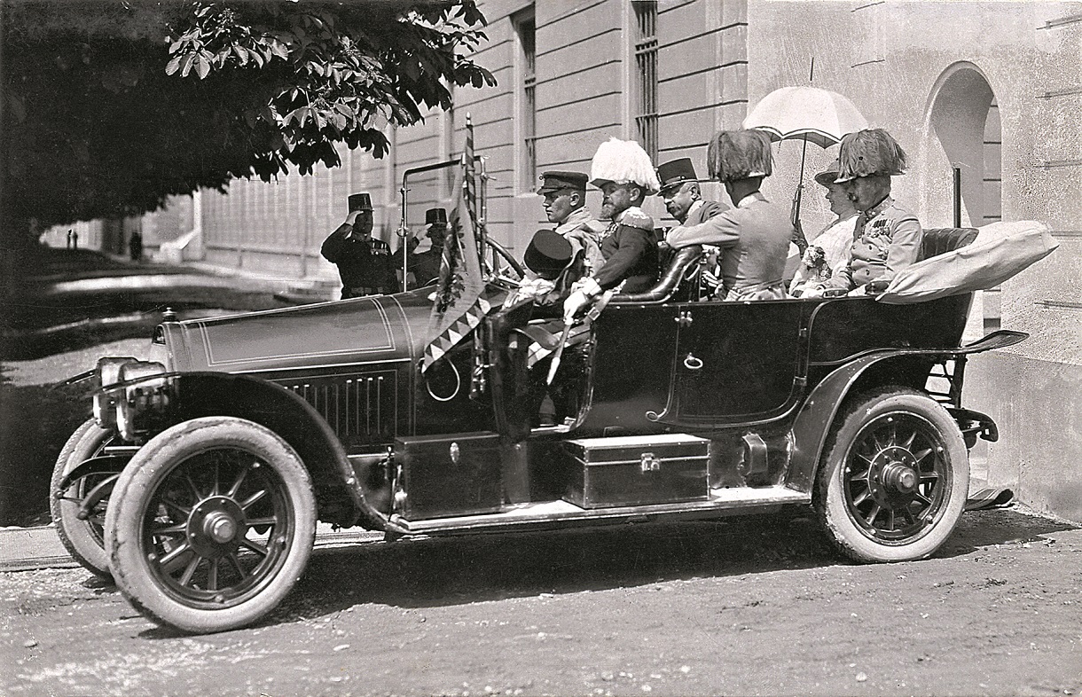 Das Schwarz-Weiß-Foto zeigt das Gräf & Stift Automobil, in dem der Thronfolger Franz Ferdinand und seine Frau Sophie Chotek sowie vier weitere Personen sitzen. Im Hintergrund salutieren Soldaten. Dieses historische Bild dokumentiert den Moment kurz vor dem ersten misslungenen Attentat, als das königliche Paar auf dem Weg zum Rathaus war