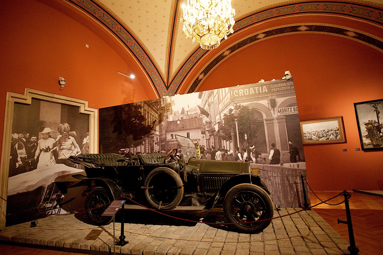 Das Bild zeigt das Automobil in seiner vollen Pracht mit offenem Verdeck, das auf einem kleinen Holzpodest steht und hinter Absperrkordeln positioniert ist. Hinter dem Fahrzeug sind Bilder zu sehen, die die letzten Momente des Paares vor dem Attentat festhalten. Die Szene wird von einem majestätischen Kronleuchter an der Decke beleuchtet, während die passend roten Wände eine stimmungsvolle Kulisse bilden.