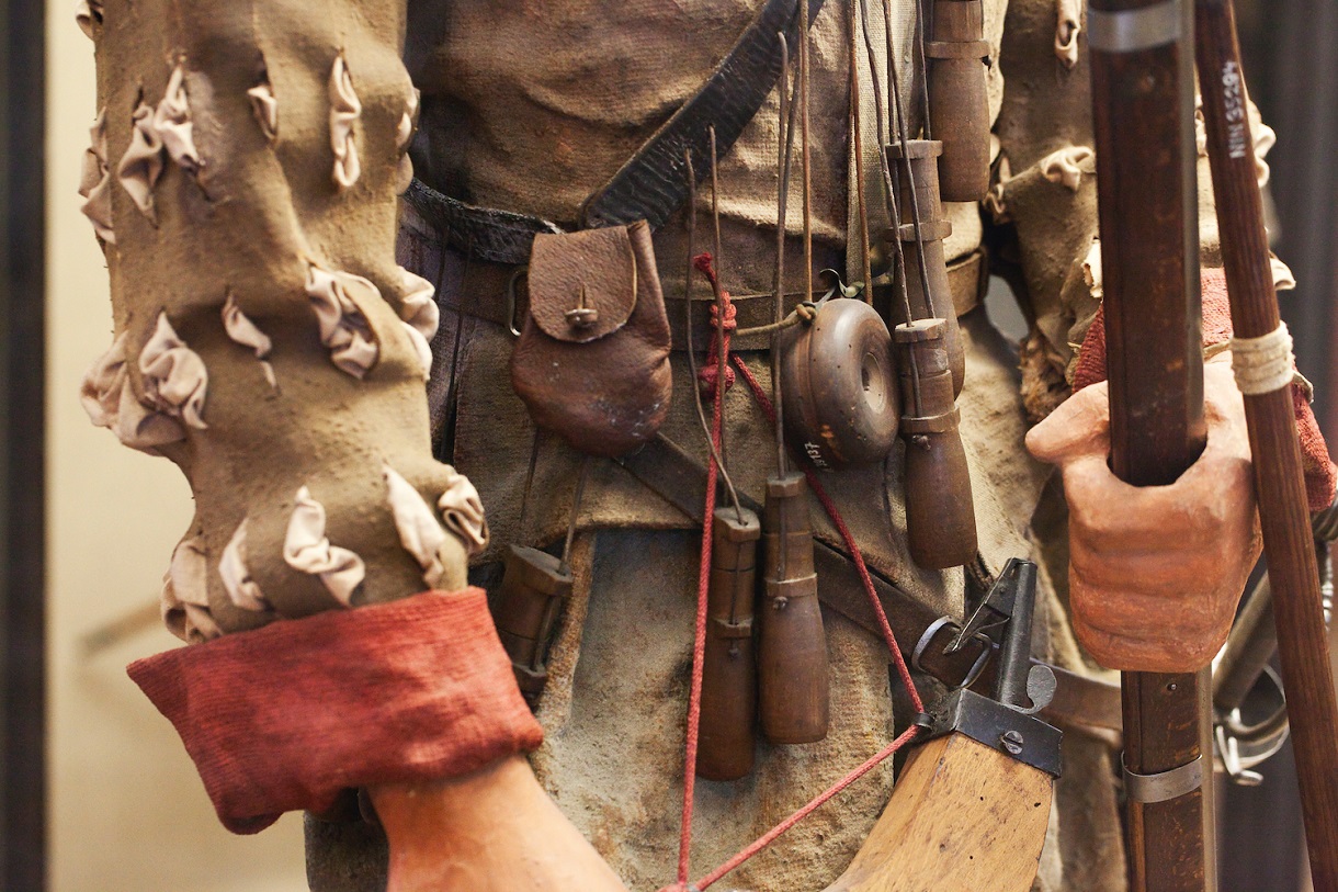  Diese Nahaufnahme präsentiert eine Puppe, die hinter einer Vitrine steht. Die hölzerne Hand der Puppe hält eine Waffe fest, während die Detailaufnahme einen faszinierenden Einblick in die fein gearbeiteten, in natürlichen Brauntönen gehaltenen Kleidungsstücke, die Munition und die Waffe selbst bietet. Jedes Element ist mit einer beeindruckenden Genauigkeit dargestellt, was die Handwerkskunst und das historische Ambiente der Ausstellung betont.