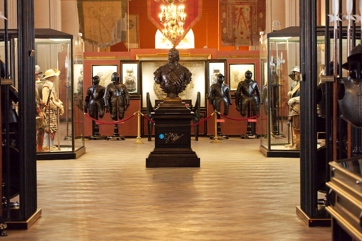 Dieses Bild zeigt eine Ausstellung mit einer braunfarbenen Statue von Prinz Eugen in der Mitte. Dahinter stehen auf jeder Seite jeweils zwei dunkelfarbene Ritterrüstungen. Neben den beiden Rüstungen befinden sich jeweils eine Vitrine mit Puppen in mittelalterlicher Kleidung in gegenüberstehender Stellung. Über der Statue hängt ein Kronleuchter, der den Raum beleuchtet. Der Boden ist hölzern und die Wände sind rot bemalt. Im Hintergrund hängen prachtvolle Wappen an der Wand.