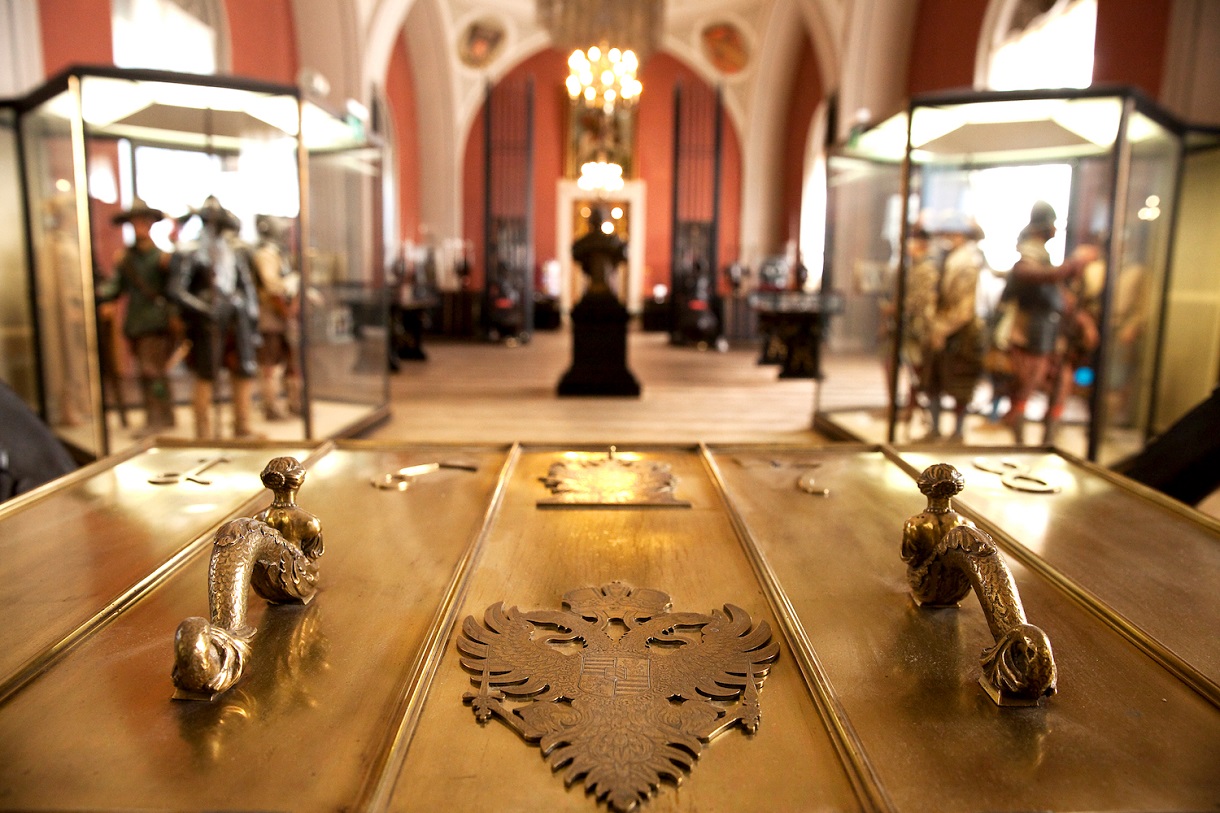 Dieses Bild zeigt die Statue von Prinz Eugen, die in der Mitte steht, während der Hintergrund verschwommen erscheint. Um die Statue herum stehen Vitrinen mit Rüstungen. In der Mitte des Bildes, direkt vor dem Auge des Betrachters, befindet sich eine goldenfarbene Truhe mit verzierten Haltegriffen. Zwischen den Griffen zeigt die Truhe das alte Wappen mit dem Doppeladler der Habsburgermonarchie. Die Detailverliebtheit der Truhe verleiht der Szene eine zusätzliche historische Tiefe.
