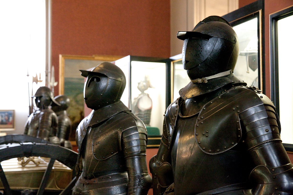 Dieses Bild zeigt vier ausgestellte Ritterrüstungen in der Dauerausstellung 'Vom Dreißigjährigen Krieg bis Prinz Eugen'. Die Rüstungen sind in dunkler Farbe gehalten. Die vorderste Ritterrüstung hat einen Helm mit einem visierartigen Deckel, der mit kleinen Löchern verziert ist, wodurch die Sicht des Ritters stark eingeschränkt war. Im Hintergrund sind weitere Rüstungen in Vitrinen zu erkennen, die jedoch aufgrund des leicht verblassten Hintergrunds schwer zu unterscheiden sind. Der Raum ist mit natürlichem Licht beleuchtet, was die Details der Rüstungen gut zur Geltung bringt.
