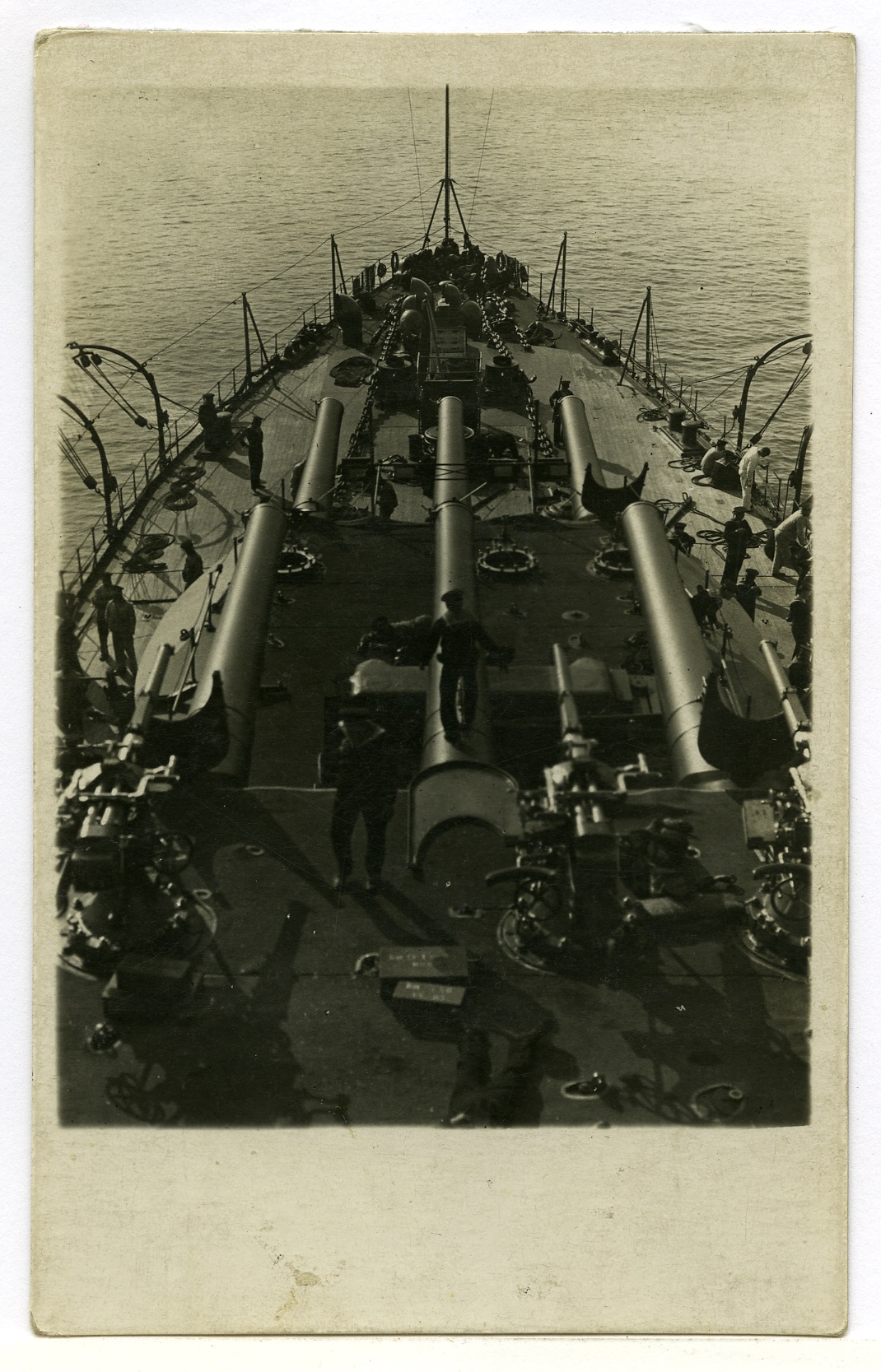 Dieses schwarz-weiß Foto zeigt einen Blick von der Brücke des Schlachtschiffes S.M.S. Viribus Unitis auf die beiden vorderen Geschütztürme, die parallel zueinander aufgestellt sind. Auf dem Deck sind einige Matrosen, die mit verschiedenen Aufgaben beschäftigt sind. Einige von ihnen stehen in der Nähe der Geschütze, andere gehen entlang der Reling. Die Uniformen und die Haltung der Besatzungsmitglieder deuten auf die Disziplin und den Alltag an Bord eines Kriegsschiffes hin. Das ruhige Meer, das das Schiff umgibt, vermittelt eine Atmosphäre von Ruhe und Stärke. 