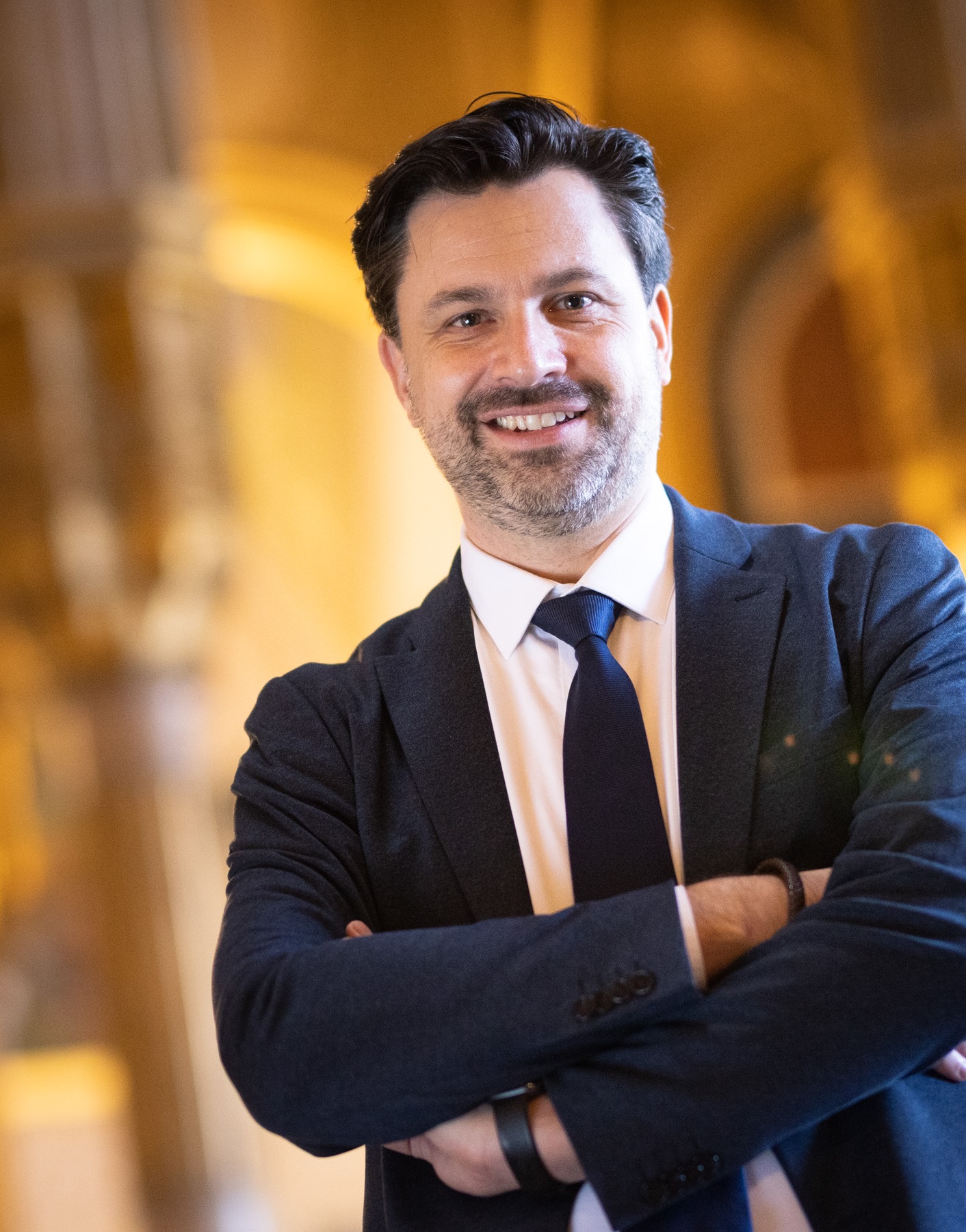 Das Bild zeigt Georg Hoffmann, den Direktor des Heeresgeschichtlichen Museums. Er steht mit verschränkten Armen und lächelt in die Kamera. Herr Hoffmann trägt einen dunkelblauen Anzug, ein weißes Hemd und eine dunkelblaue Krawatte. Sein Haar ist dunkel und leicht zurückgekämmt, und er hat einen gepflegten Bart. Der Hintergrund ist unscharf, aber es lässt sich erkennen, dass er sich in einem prächtigen, gut beleuchteten Raum mit architektonischen Details befindet, was auf eine historische oder offizielle Umgebung hinweist. Herr Hoffmann wirkt freundlich, selbstbewusst und professionell.