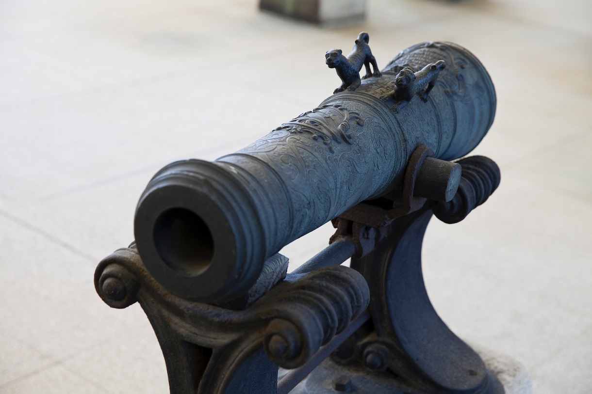 Auf dem Bild ist ein kleines Geschützrohr zu sehen, das durch seine kunstvollen Verzierungen und verschnörkelten Standbeine auffällt. Das Geschützrohr selbst zeigt auf seiner Oberfläche aufwendige Gravuren und Muster, sowie ein Wappen, die meisterhaft eingearbeitet wurden. Besonders ins Auge fallen zwei tierische Wesen, die kunstvoll an das Rohr integriert sind. 