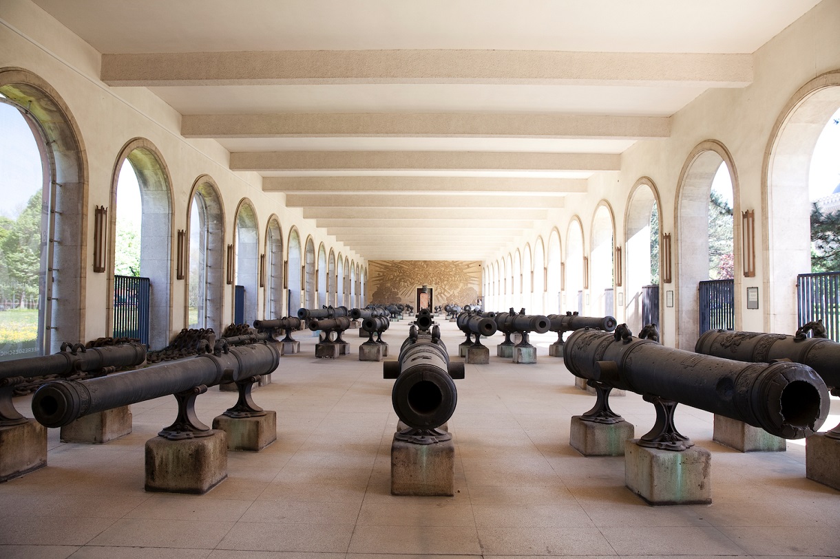  Auf dem Bild ist die beeindruckende Artilleriehalle zu sehen, in der verschiedene historische Geschütze ausgestellt sind. Die Wände sowie der Boden der Halle sind schlicht gehalten, die Halle ist geräumig und durch die umliegenden Bögen gut beleuchtet, was die Exponate ins rechte Licht rückt. Im Hintergrund ist eine Wand mit einer künstlerischen Kriegsdarstellung in heller Farbe zu erkennen, das historische Szenen darstellt und der Halle eine besondere Atmosphäre verleiht. Die Geschützrohre variieren in Größe und Form und repräsentieren verschiedene Epochen der Artilleriegeschichte. Diese Vielfalt zeigt die handwerkliche und technologische Entwicklung der Artillerie über die Jahrhunderte hinweg. 