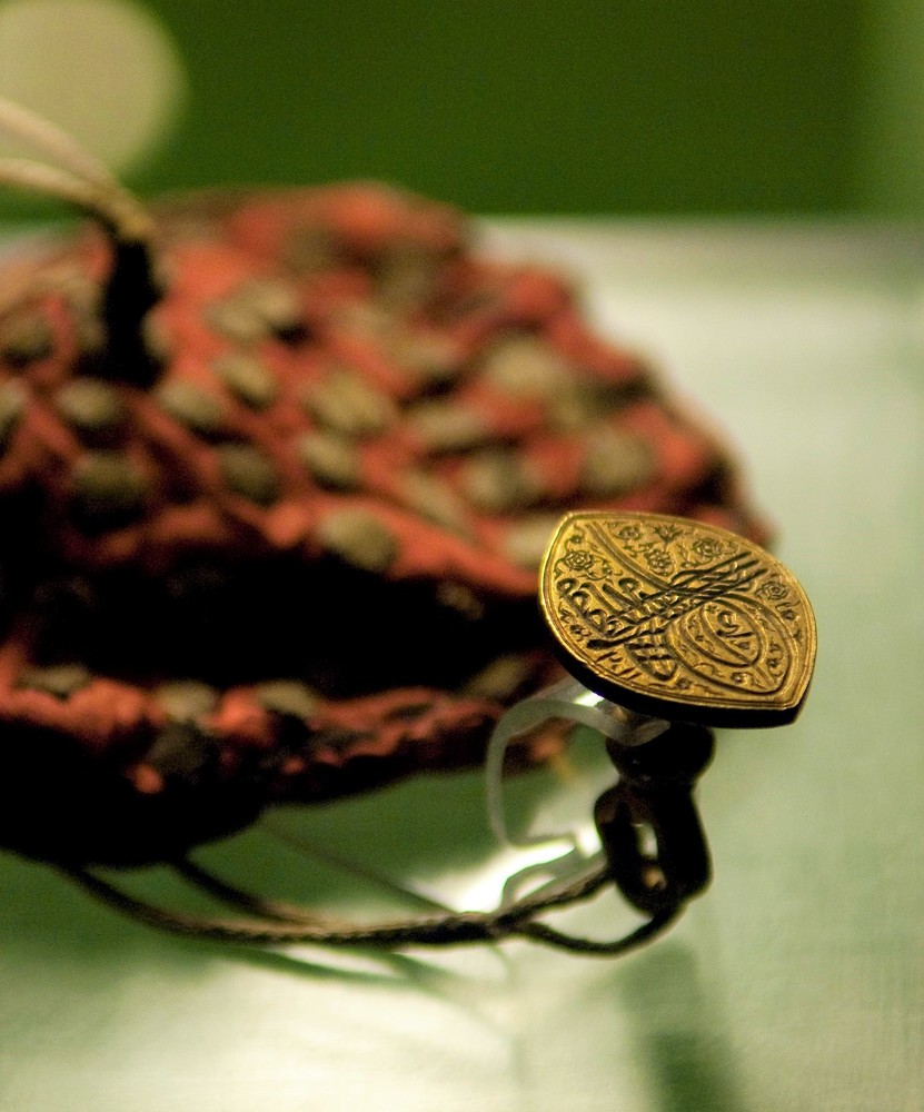 Diese Nahaufnahme zeigt einen historischen Beutel als Ausstellungsobjekt, der auf einer Oberfläche in hellgrünen Tönen liegt. Der Beutel selbst ist in Rot- und Brauntönen gestaltet und strahlt eine warme und einladende Ästhetik aus. An ihm hängt ein münzgroßer goldener Anhänger mit kunstvollen Verzierungen und einer Inschrift. Der Anhänger zeigt das Siegel eines türkischen Sultans, was auf seine historische Bedeutung hinweist. Die feinen Details des Anhängers, einschließlich der kunstvollen Verzierungen und der Inschrift, sind deutlich erkennbar.