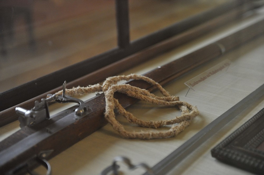 Dieses Bild zeigt eine Luntenmuskete hinter einer Glasvitrine. Die Muskete ist detailreich gearbeitet und gut erhalten. Die Vitrine schützt die historische Waffe und ermöglicht eine klare Sicht auf die Details der Mechanik und des Holzschafts. Der Raum ist gut beleuchtet, wodurch die Besonderheiten der Luntenmuskete deutlich hervortreten.