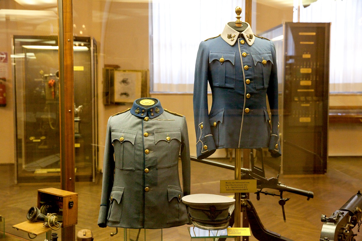 Das Bild zeigt eine Vitrine mit zwei Uniformen des Habsburgerreiches, einem Feldtelefon, einem Hut und verschiedenen Waffen. Diese Ausstellungsstücke bieten einen Einblick in die Militärgeschichte des Habsburgerreiches und seine militärische Ausrüstung. 