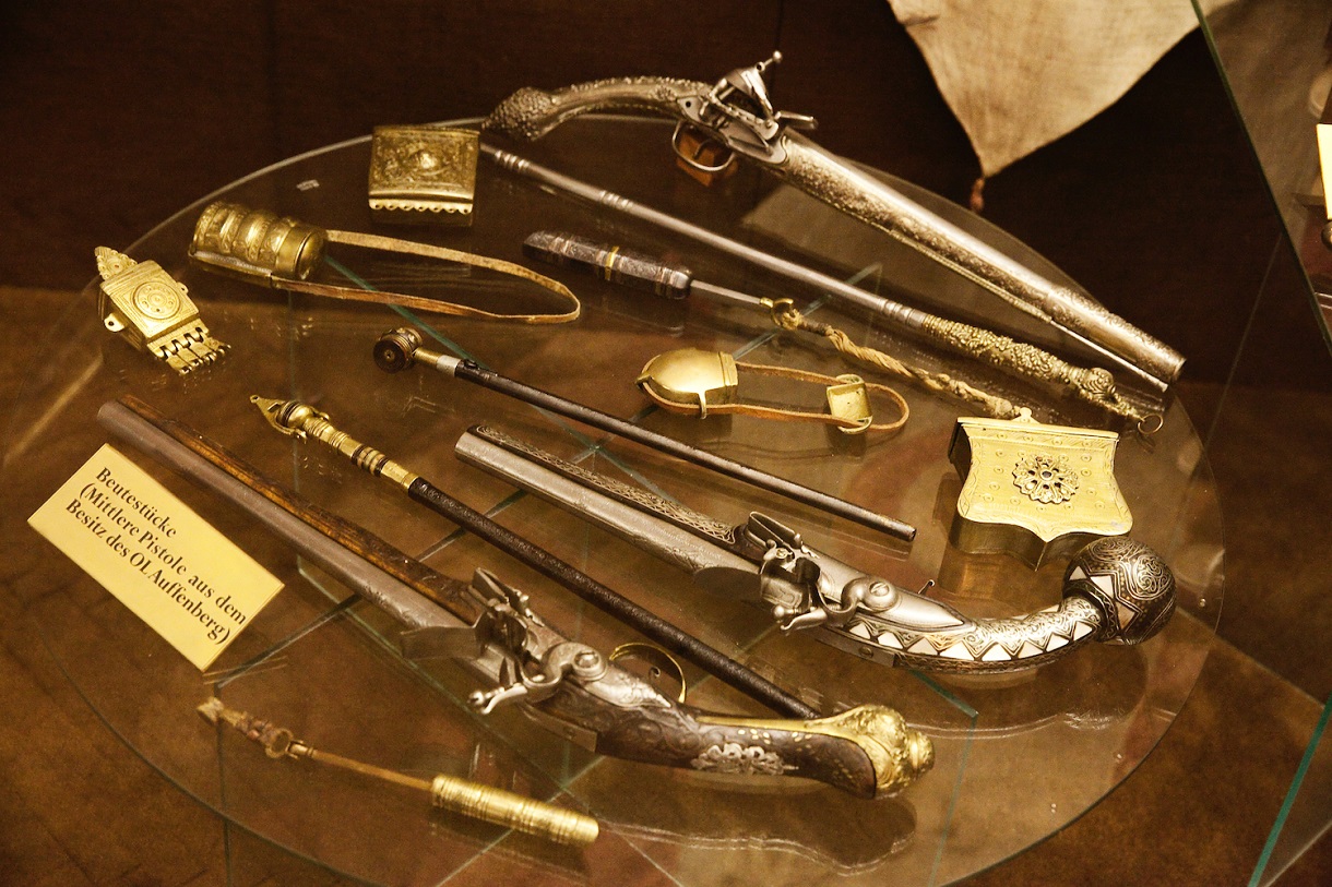 Das Bild zeigt verschiedene Handfeuerwaffen auf einem Glastisch hinter einer Vitrine, die als Beutestücke präsentiert werden. Eine der Pistolen sticht besonders hervor, da sie aus dem Besitz des OL Auffenberg stammt. 