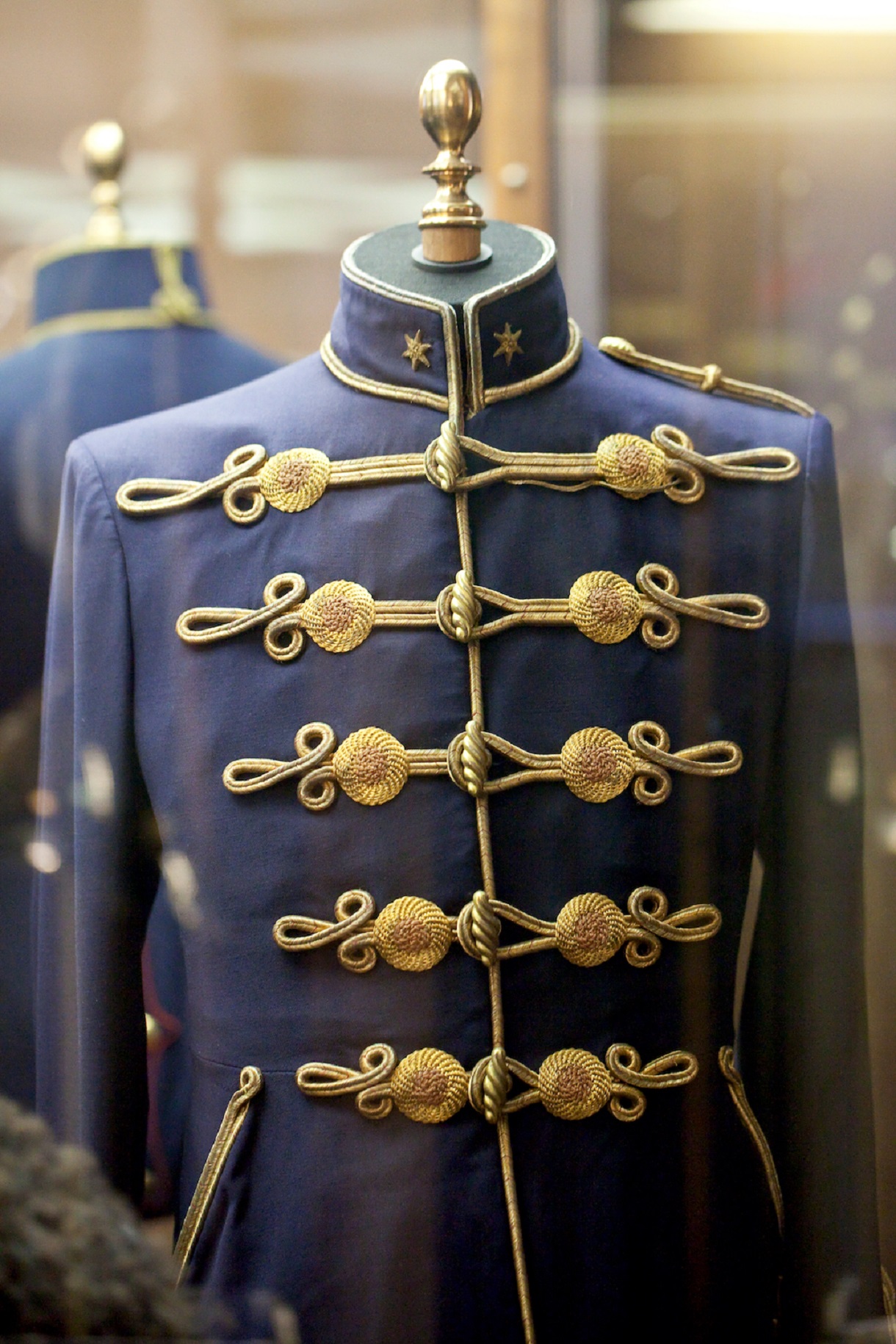 Das Bild zeigt eine Uniformjacke mit goldenen Details, die in einer Vitrine ausgestellt ist. Diese eindrucksvolle Uniform repräsentiert möglicherweise eine hochrangige militärische oder zeremonielle Position. Die goldenen Verzierungen verleihen der Jacke eine herausragende Pracht und symbolisieren Würde und Autorität. 