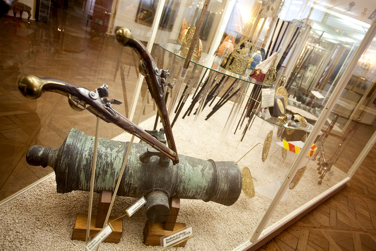 Dieses Bild bietet einen Einblick in die Zeit des 18. Jahrhunderts durch die Ausstellung einer Kanone und zwei Musketen in einer Vitrine. Die grünliche Verfärbung der Kanone, vermutlich aufgrund der Witterung, sowie die golden verzierten Griffe der Musketen vermitteln die Materialität und Ästhetik dieser Epoche. Diese Waffenrepräsentationen ermöglichen es den Betrachtern, sich ein Bild von den Technologien und dem Stil dieser Zeit zu machen und bieten somit einen Einblick in die militärische Geschichte des 18. Jahrhunderts.