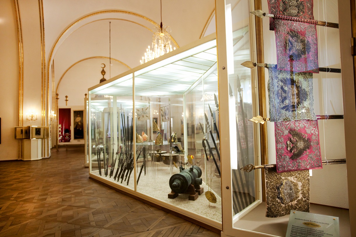 Dieses Bild bietet einen Einblick in die Zeit des 18. Jahrhunderts durch die Ausstellung einer Kanone und zwei Musketen in einer Vitrine. Die grünliche Verfärbung der Kanone, vermutlich aufgrund der Witterung, sowie die golden verzierten Griffe der Musketen vermitteln die Materialität und Ästhetik dieser Epoche. Vor der Vitrine hängen verschiedenfarbige Flaggen, ebenfalls in einer Vitrine gehalten, die die Vielfalt der Nationen und Kulturen der damaligen Zeit repräsentieren. Der Raum ist mit einem prunkvollen Kronleuchter beleuchtet, der eine königliche Atmosphäre schafft und die Bedeutung der ausgestellten Artefakte unterstreicht.