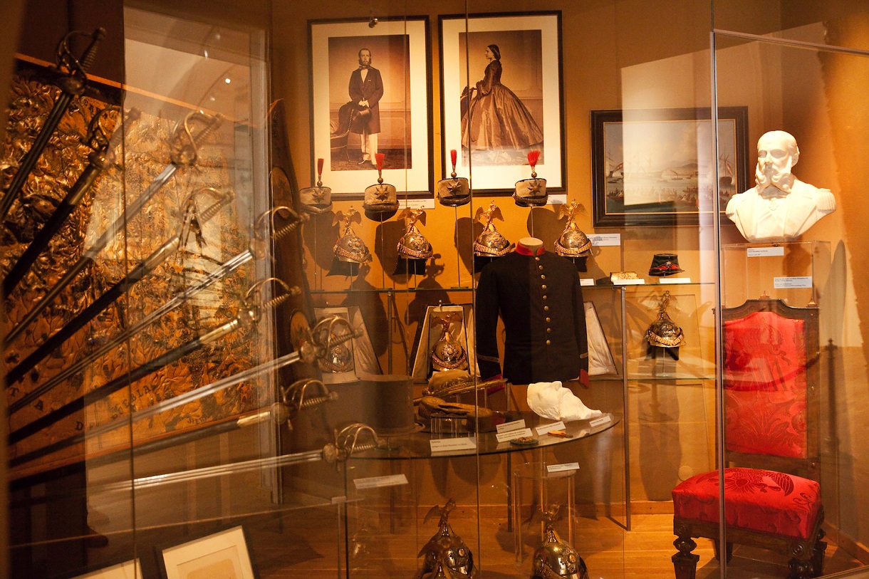 Das Bild zeigt eine Vitrine mit Schwertern, dahinter ausgestellte Helme, Uniformjacken und Gemälde. Im Hintergrund hängen schwarz-weiß Fotos des Kaiserpaares. Diese Artefakte sind Teil der Ausstellung über Maximilians Kaiserreich in Mexiko. 