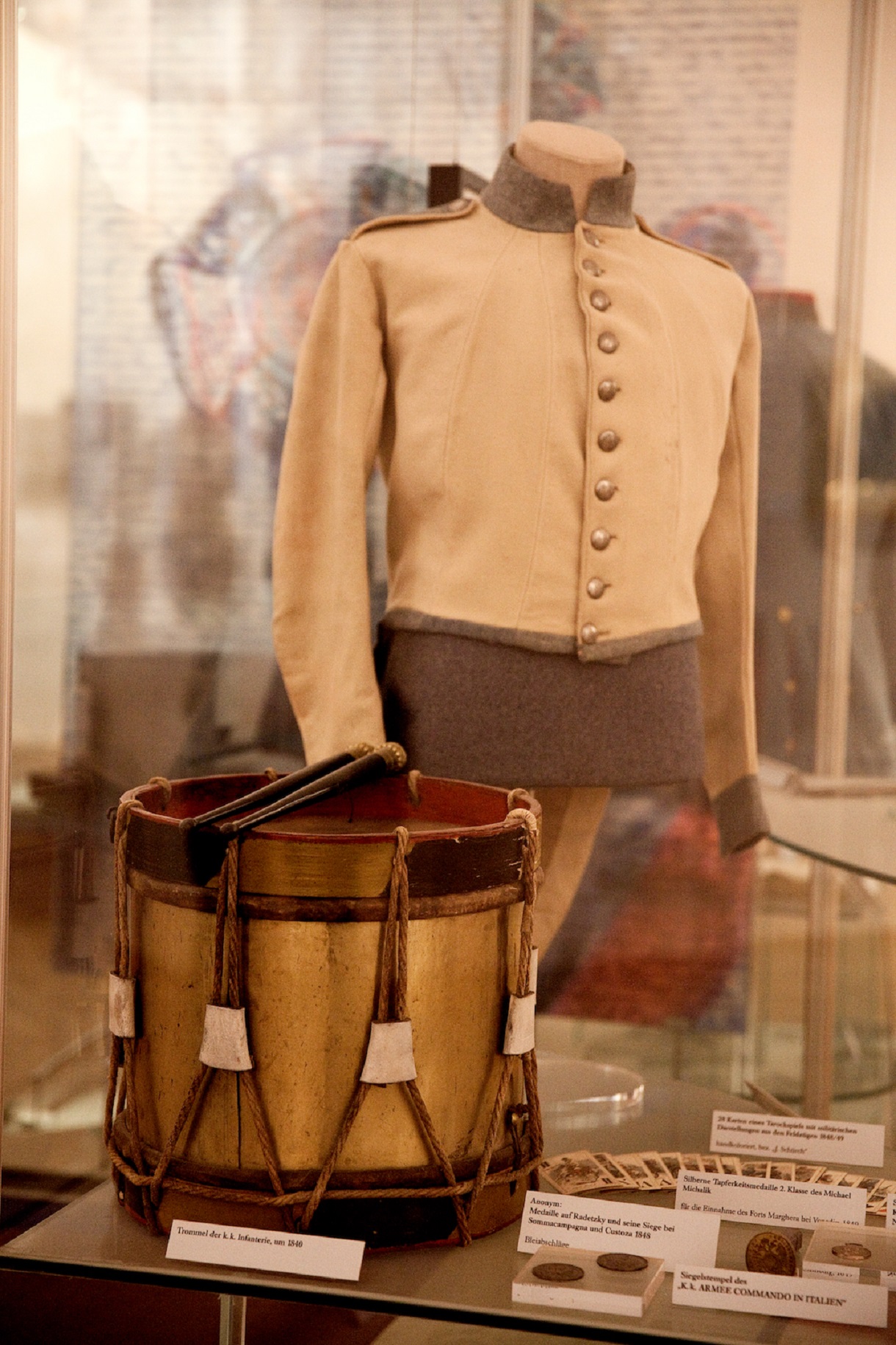Das Bild zeigt eine Holztrommel neben Medaillen aus Blei und Silber, mit einer Uniformjacke im Hintergrund, alle in einer Vitrine ausgestellt. Ihre Präsentation bietet einen Einblick in die militärische Geschichte und Tradition.