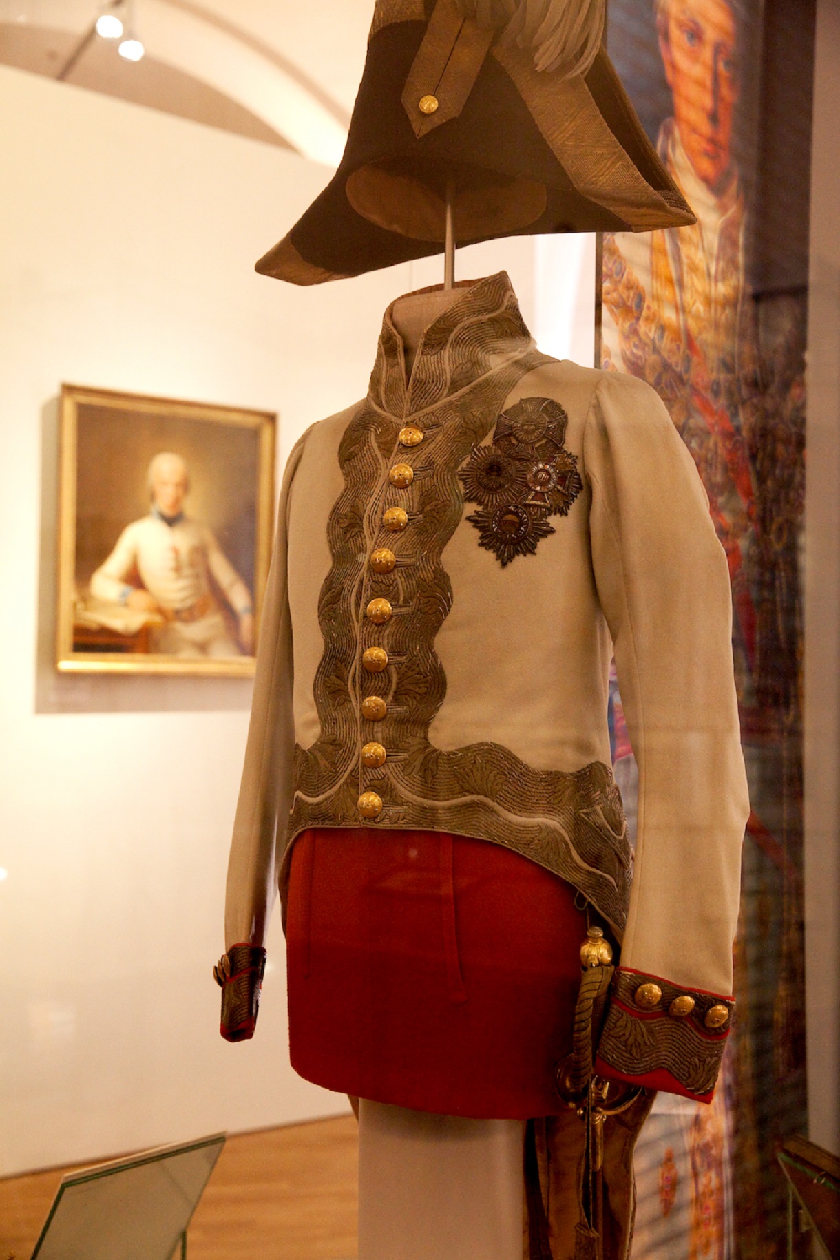  Das Bild zeigt die Gala-Uniform von Kaiser Franz I., bestehend aus einer roten Hose und einem weißen Jacket mit goldenen Knöpfen. Die Uniform wird in einer Vitrine aufbewahrt, die ihre Pracht betont. Im Hintergrund ist ein unscharfes Gemälde eines Selbstporträts zu erkennen, dessen vergoldeter Rahmen den königlichen Glanz und die Pracht der damaligen Zeit widerspiegelt.