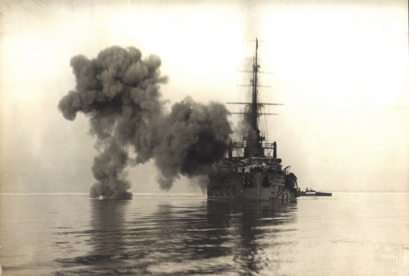 Ein dramatisches Schwarz-Weiß-Foto zeigt die SMS Viribus Unitis, ein mächtiges Schlachtschiff, die ihre mächtigen Geschütze abfeuert. Dichte Rauchwolken steigen von den Kanonen auf und hüllen das Schlachtschiff in eine dramatische Szenerie. Die raue Textur der Wellen und die Struktur des Schiffes ist klar erkennbar und die aufsteigenden Rauchschwaden verleihen dem Bild eine intensive Atmosphäre.