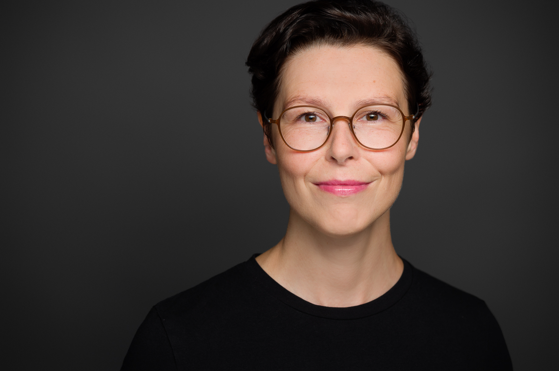 Auf dem Foto ist die Militär- und Medizinhistorikerin Sabine Jesner zu sehen. Sie hat kurzes, dunkles Haar und trägt eine runde Brille mit einem hellen Rahmen. Sie trägt ein schwarzes Oberteil und lächelt leicht in die Kamera. Der Hintergrund ist einfarbig dunkelgrau, wodurch ihr Gesicht deutlich hervorsticht. Das Porträt ist professionell aufgenommen und zeigt sie in einer ruhigen und selbstbewussten Pose.